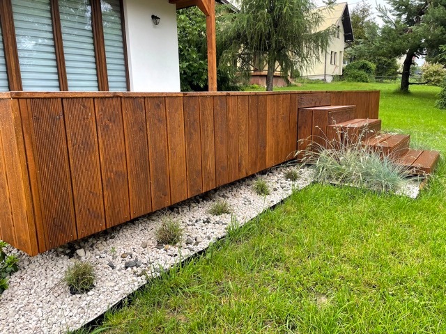 Nowoczesny taras z drewna o ciepłym odcieniu brązu, z niską zabudową i schodami, otoczony białym żwirem i zielonym trawnikiem, w tle dom z białymi ścianami i dużymi oknami.