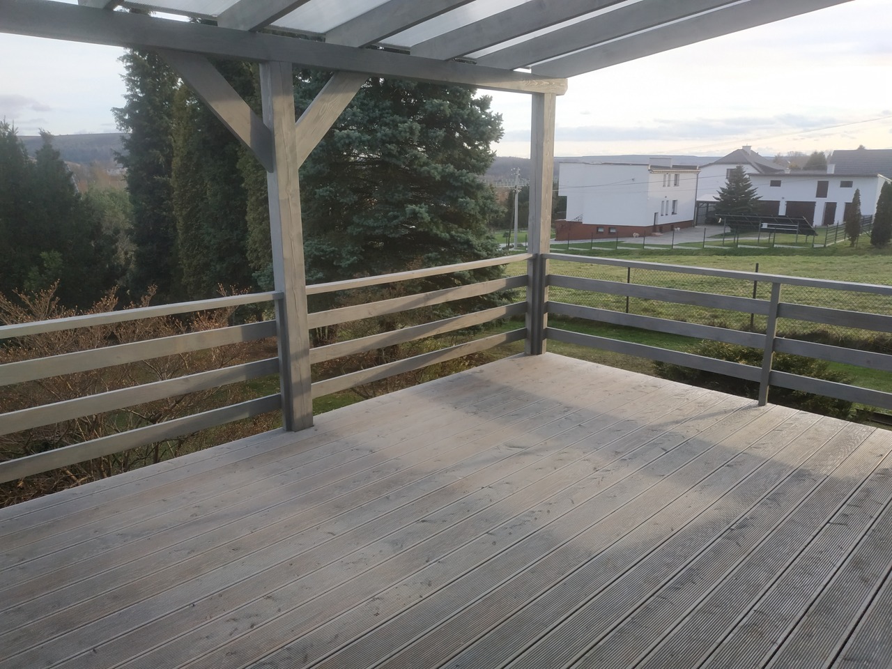 Zadaszony taras z szarych desek kompozytowych, z drewnianą balustradą w tym samym kolorze, widok na zielony ogród i zabudowania w tle.