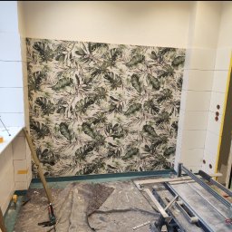 Mateusz Zając - Ściana łazienki wyłożona białymi płytkami i dekoracyjnymi płytkami z motywem tropikalnych liści, widoczny poziomicę, butelkę wody, maszynkę do cięcia płytek i inne narzędzia na podłodze...