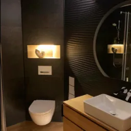 Nowoczesna łazienka z czarnymi płytkami o falistej strukturze, drewnianą szafką pod umywalką i podświetlaną niszą z dekoracją w kształcie muszli.