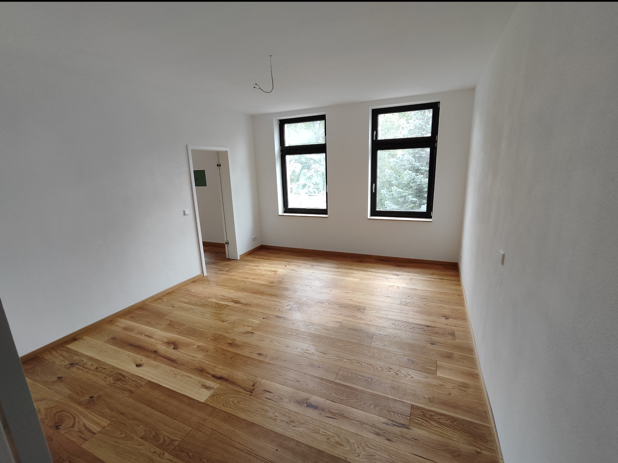 Pusty pokój po remoncie z drewnianą podłogą w odcieniach brązu i beżu, świeżo pomalowane białe ściany, dwa okna z czarnymi ramami i widokiem na zieleń za oknem, oraz widoczne zwisające kable...