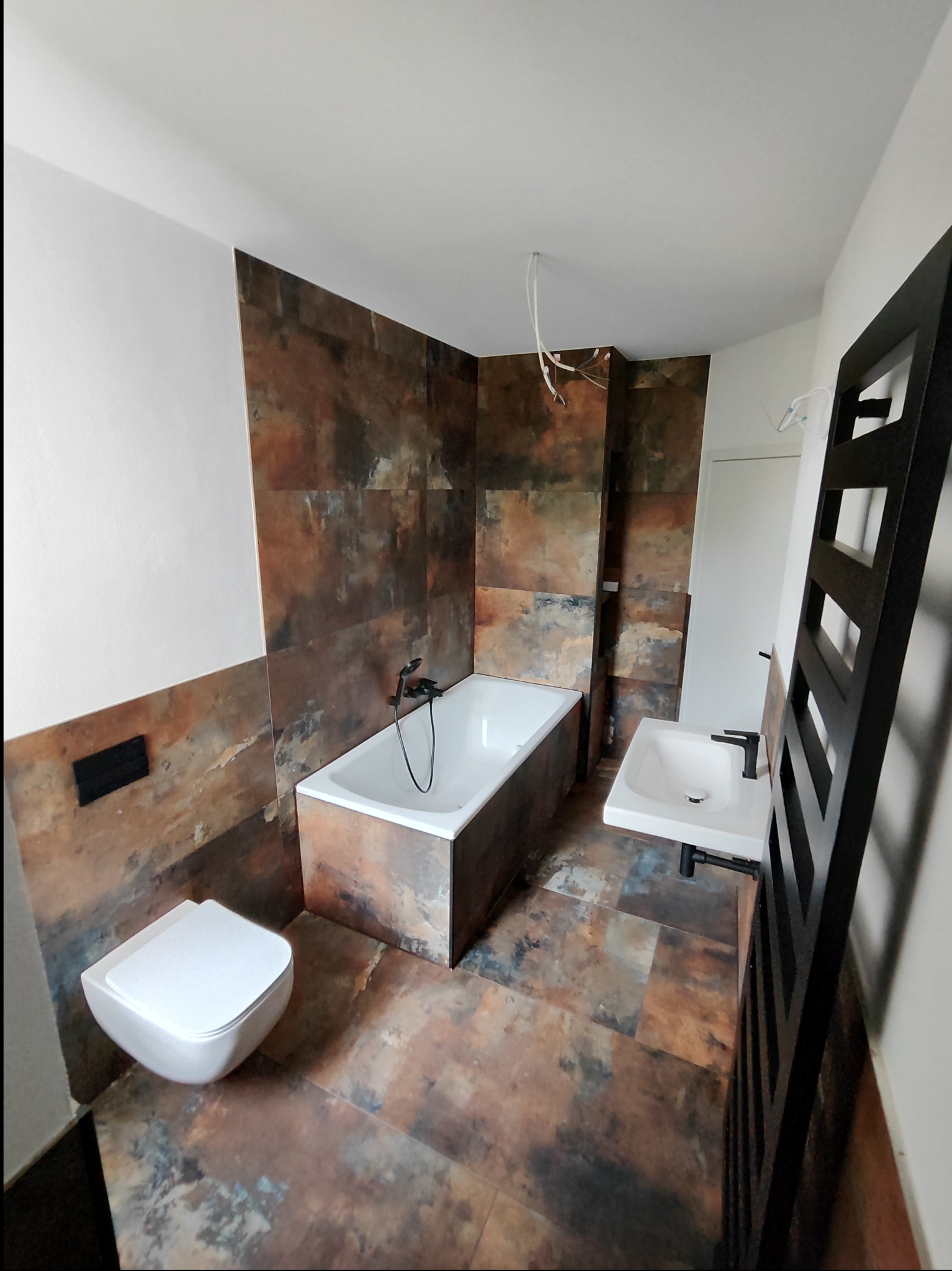 Nowoczesna łazienka z płytkami imitującymi rdzę, wisząca toaleta, biała wanna i umywalka z czarną armaturą, grzejnik drabinkowy w czarnym kolorze, surowe przewody elektryczne zwisające z sufitu.