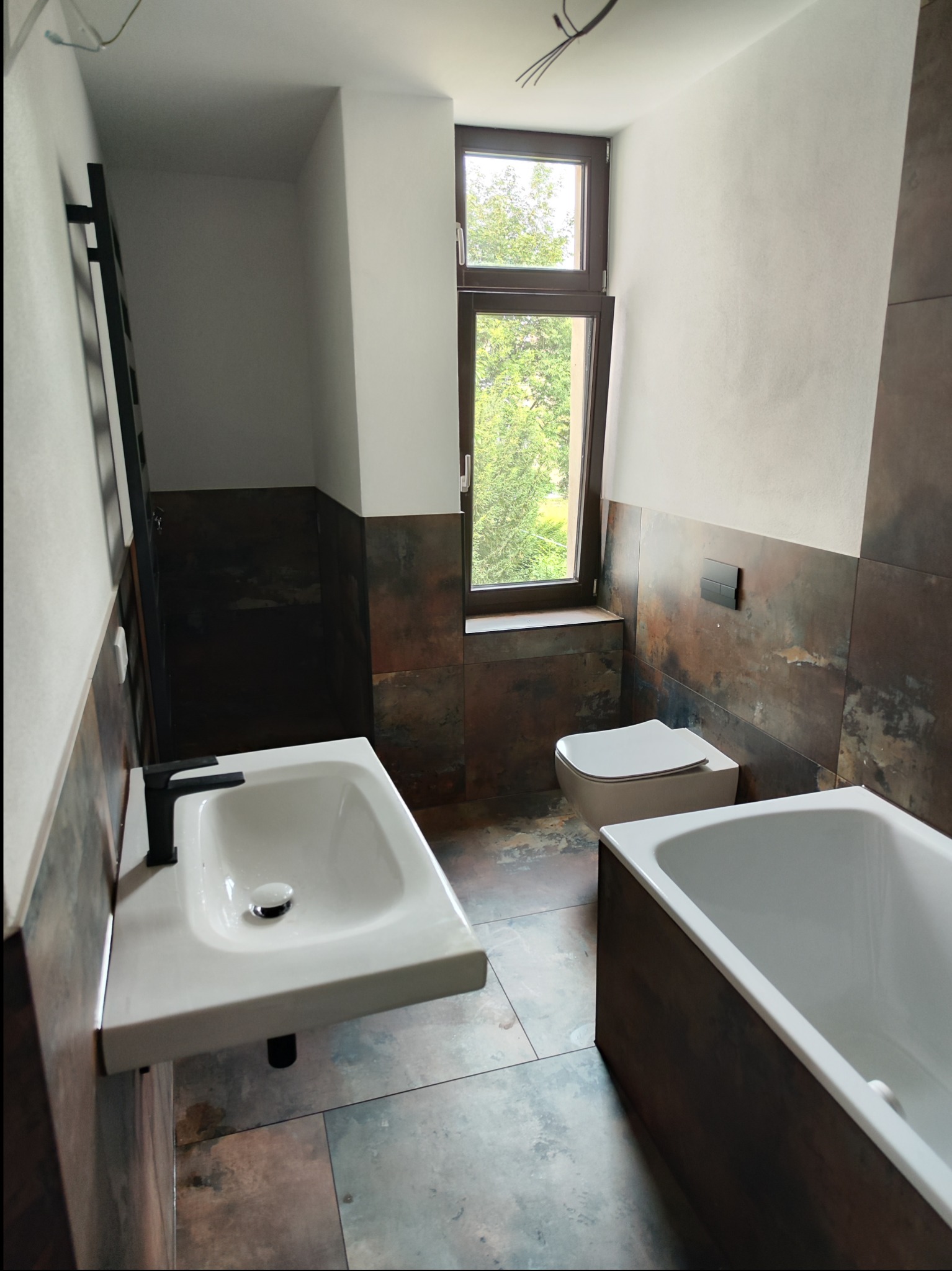 Nowoczesna łazienka z brązowymi płytkami imitującymi rdzę, białą armaturą, czarną baterią umywalkową i grzejnikiem drabinkowym, widok na okno z zielenią.