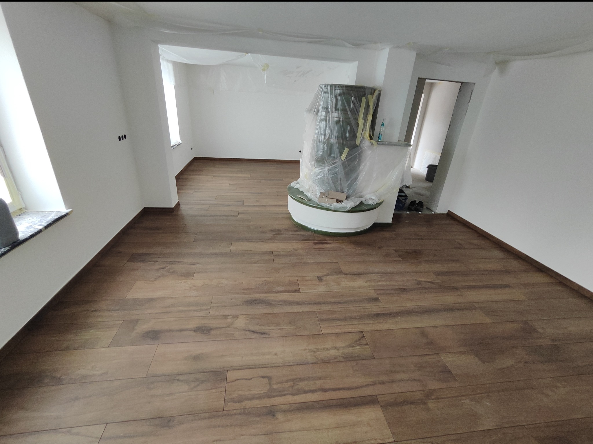 Pokój po remoncie z drewnopodobną podłogą, świeżo pomalowanymi białymi ścianami i zabezpieczonym folią piecem kaflowym.