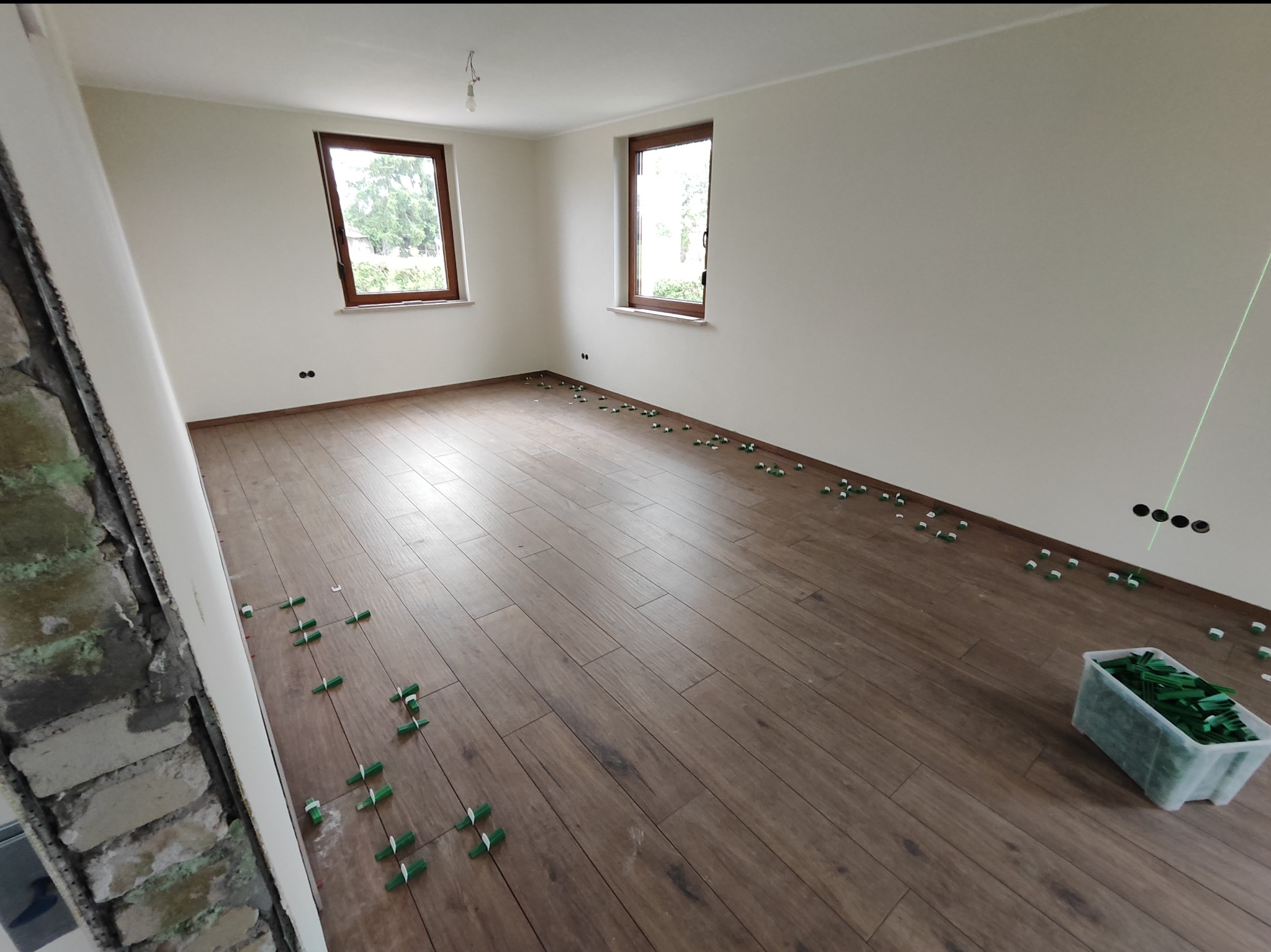 Układanie paneli podłogowych imitujących drewno, z użyciem zielonych krzyżyków dystansowych; widoczne ślady po instalacji elektrycznej i surowa ściana w tle.