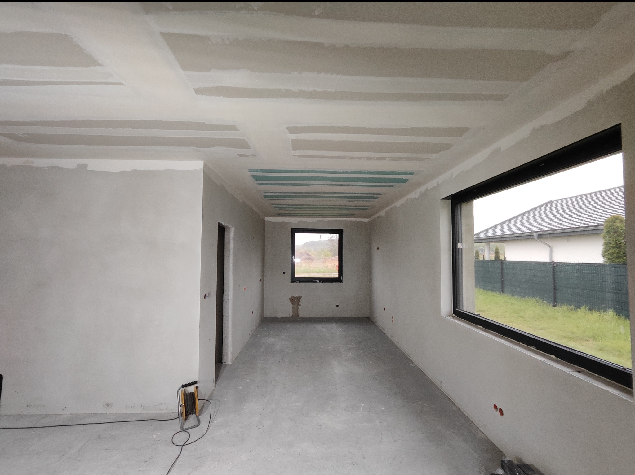 Wnętrze w trakcie prac: surowy stan wykończenia, widoczne płyty gipsowo-kartonowe na suficie przed szpachlowaniem, ściany w stanie surowym, okna z czarnymi ramami, na podłodze wylewka, zwinięty...