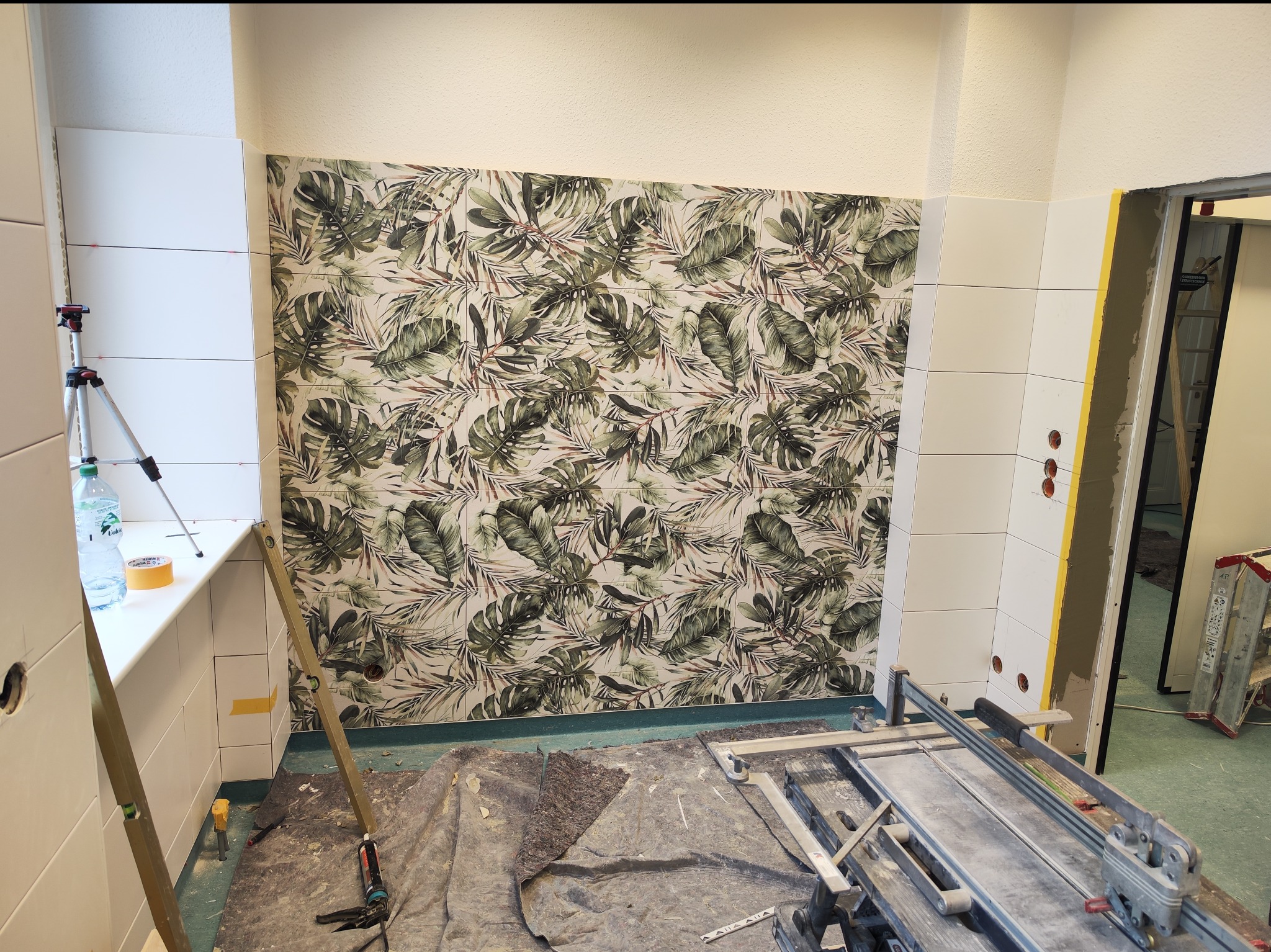 Ściana łazienki wyłożona białymi płytkami i dekoracyjnymi płytkami z motywem tropikalnych liści, widoczny poziomicę, butelkę wody, maszynkę do cięcia płytek i inne narzędzia na podłodze...