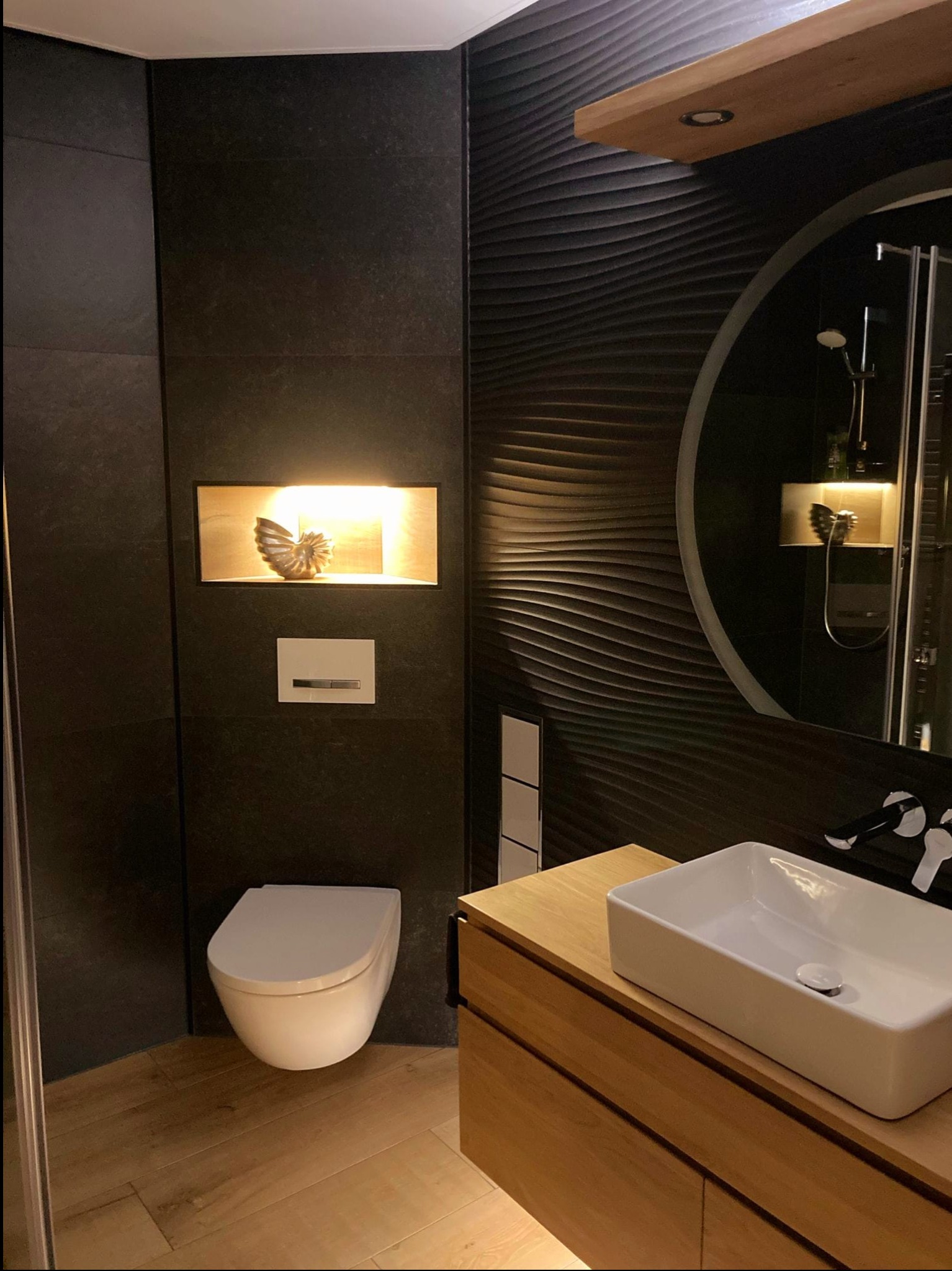 Nowoczesna łazienka z czarnymi płytkami o falistej strukturze, drewnianą szafką pod umywalką i podświetlaną niszą z dekoracją w kształcie muszli.