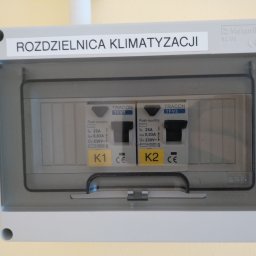 EMC Elektromechanika Chłodnicza - Szara skrzynka rozdzielcza klimatyzacji z dwoma bezpiecznikami TRACON TFV2 oznaczonymi K1 i K2, z naklejką informacyjną o parametrach elektrycznych.