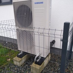 EMC Elektromechanika Chłodnicza - Zewnętrzna jednostka klimatyzacji Samsung zamontowana na betonowych bloczkach, otoczona metalowym ogrodzeniem panelowym, z widocznymi przewodami i rurą odprowadzającą skropliny.