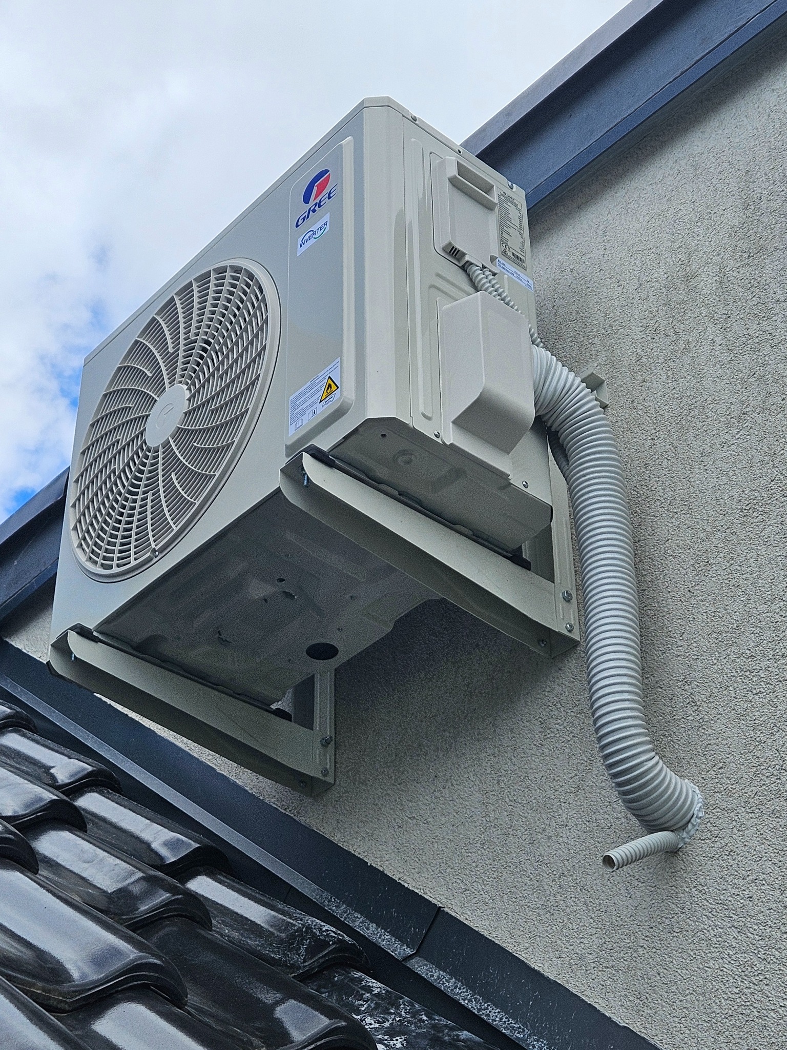 Zewnętrzna jednostka klimatyzatora marki Gree zamontowana na ścianie budynku z widocznym przewodem odprowadzającym skropliny, zainstalowana nad fragmentem dachu pokrytego błyszczącą dachówką.