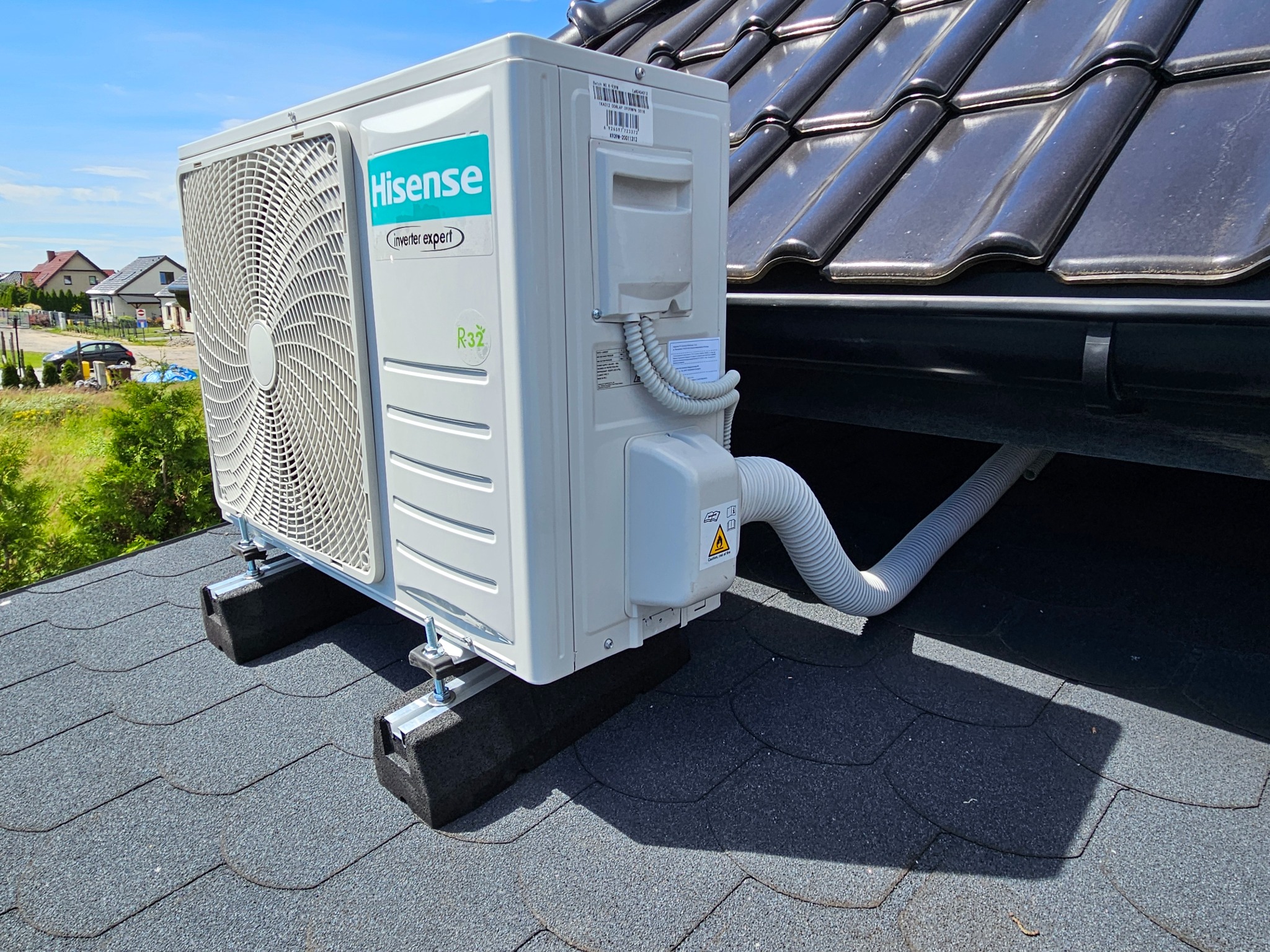 Zewnętrzna jednostka klimatyzacji Hisense zamontowana na dachu pokrytym gontem bitumicznym, widoczne czarne podkładki tłumiące drgania i elementy mocujące.