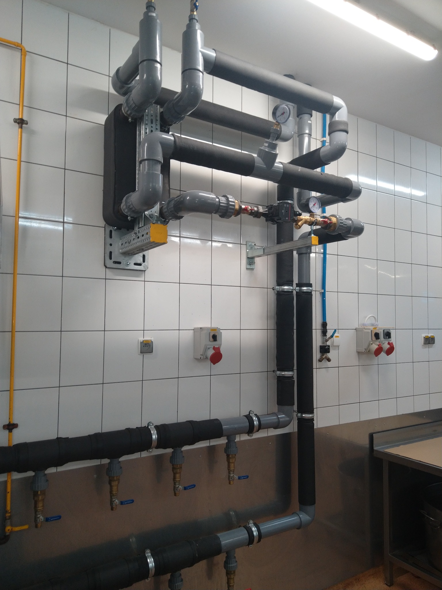 Rozbudowany system rur z izolacją termiczną i manometrami zamontowany na ścianie z białych płytek w pomieszczeniu przemysłowym.