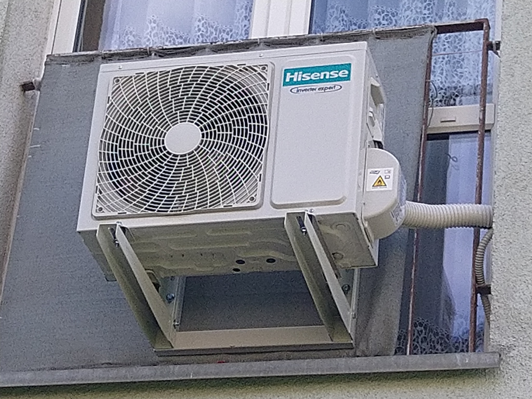 Zewnętrzna jednostka klimatyzacji Hisense zamontowana na balkonie, widoczna kratka wentylacyjna i rura odprowadzająca skropliny.