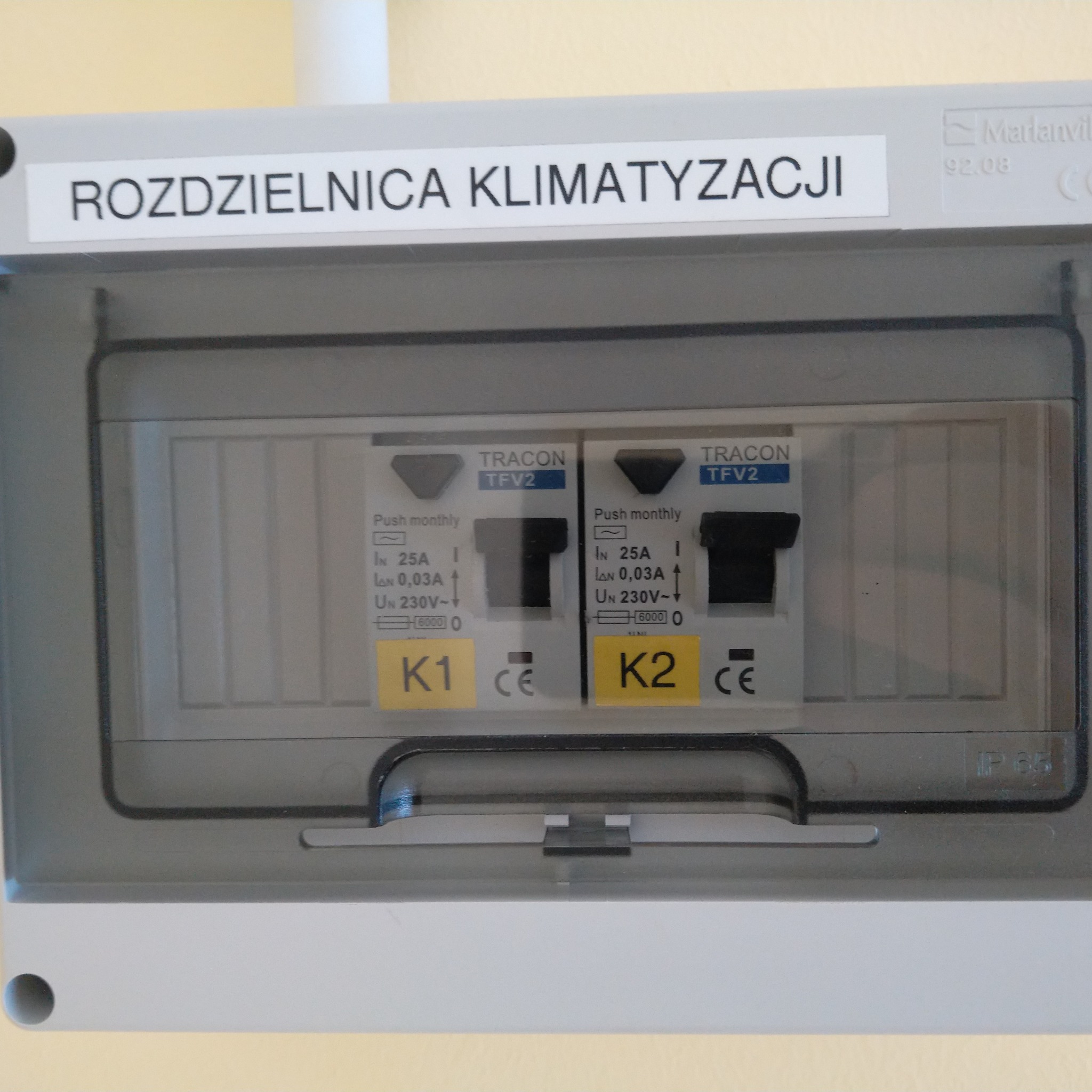 Szara skrzynka rozdzielcza klimatyzacji z dwoma bezpiecznikami TRACON TFV2 oznaczonymi K1 i K2, z naklejką informacyjną o parametrach elektrycznych.