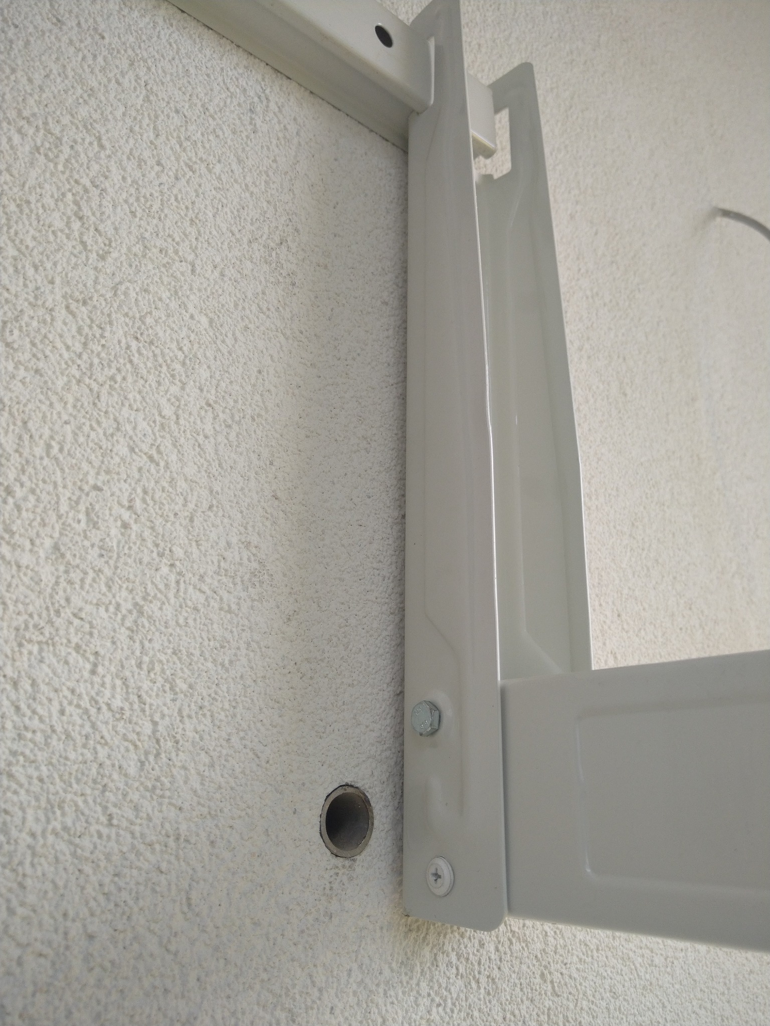 Biały, metalowy wspornik klimatyzatora zamontowany na szorstkiej, białej ścianie. Widoczny wywiercony otwór obok wspornika.