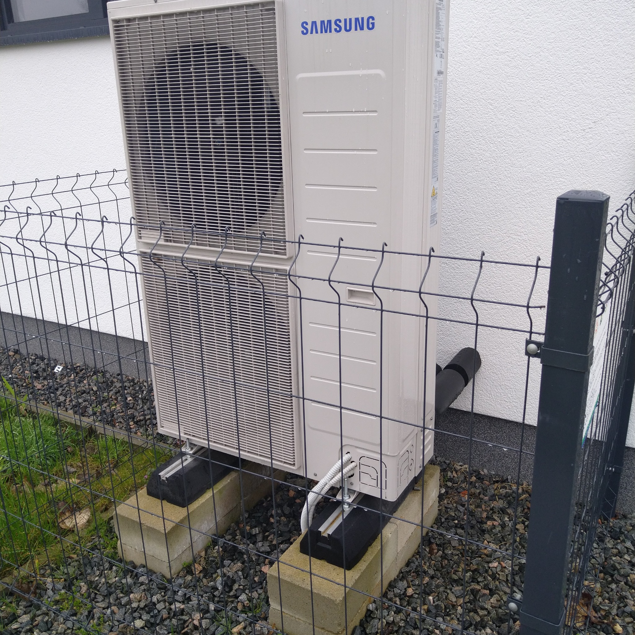 Zewnętrzna jednostka klimatyzacji Samsung zamontowana na betonowych bloczkach, otoczona metalowym ogrodzeniem panelowym, z widocznymi przewodami i rurą odprowadzającą skropliny.