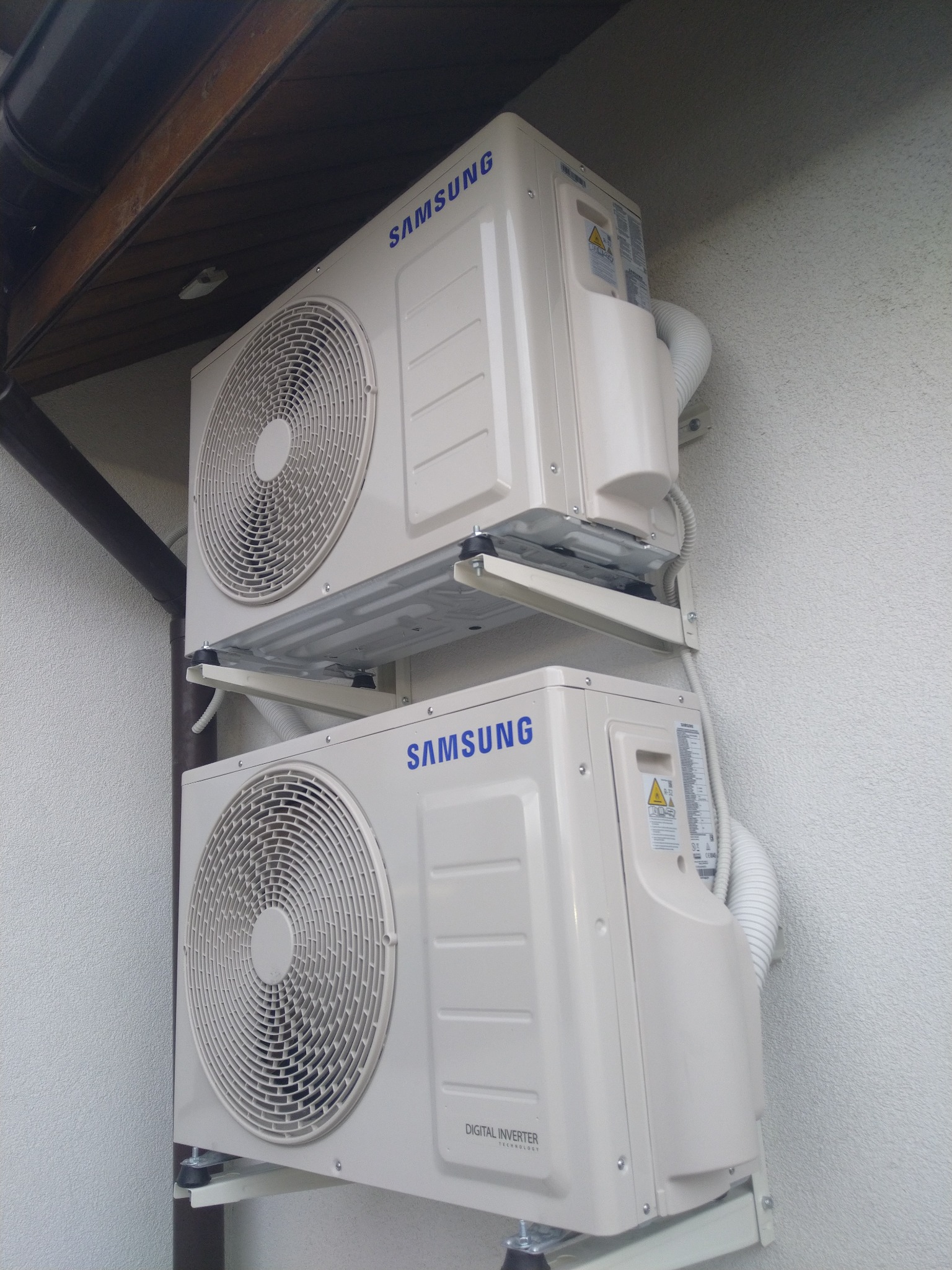 Dwa zewnętrzne moduły klimatyzacji Samsung Digital Inverter zamontowane jeden nad drugim na wspornikach na jasnej ścianie budynku.