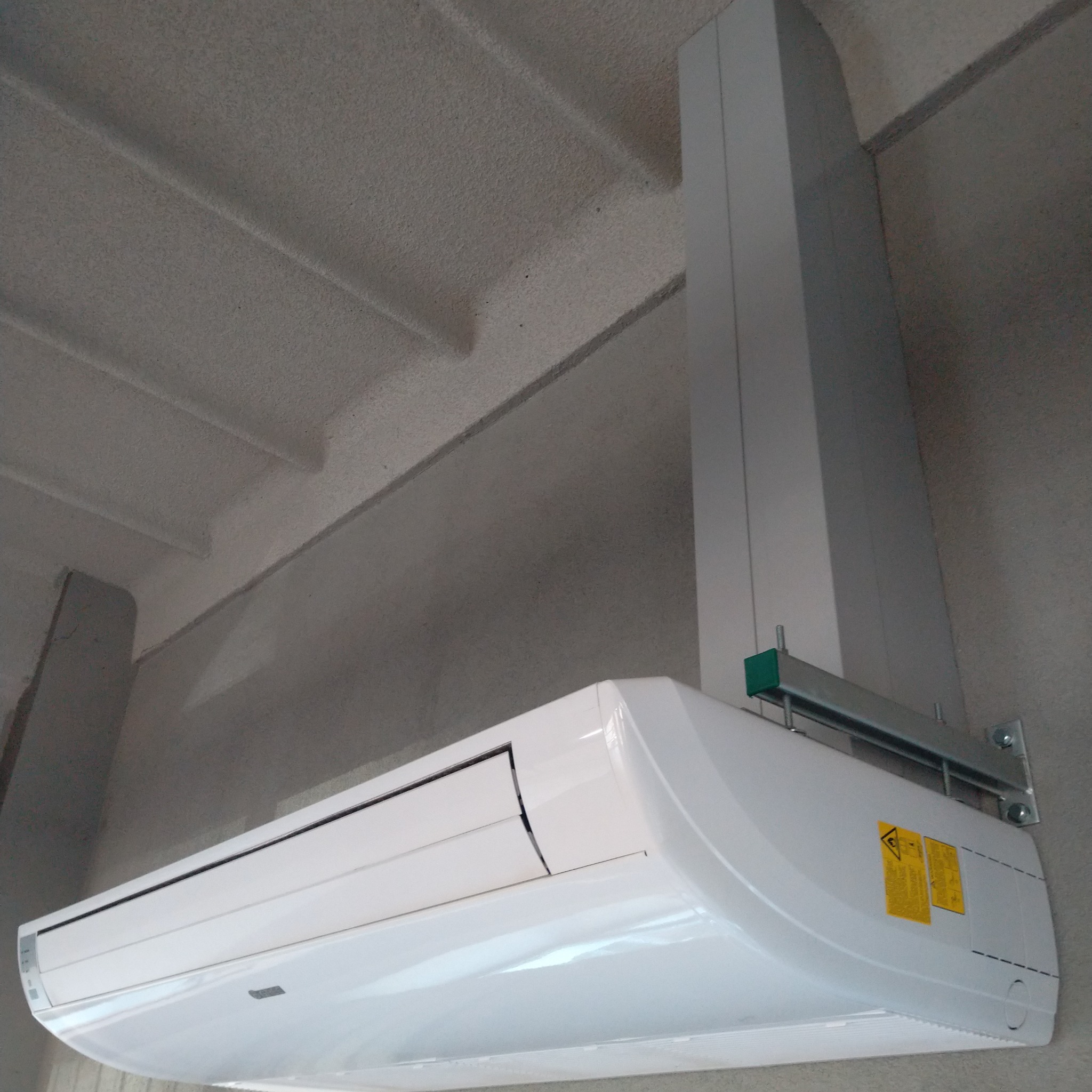 Biała jednostka wewnętrzna klimatyzacji zamontowana na ścianie z widocznymi elementami mocującymi i kanałem maskującym instalację elektryczną na tle surowego sufitu.