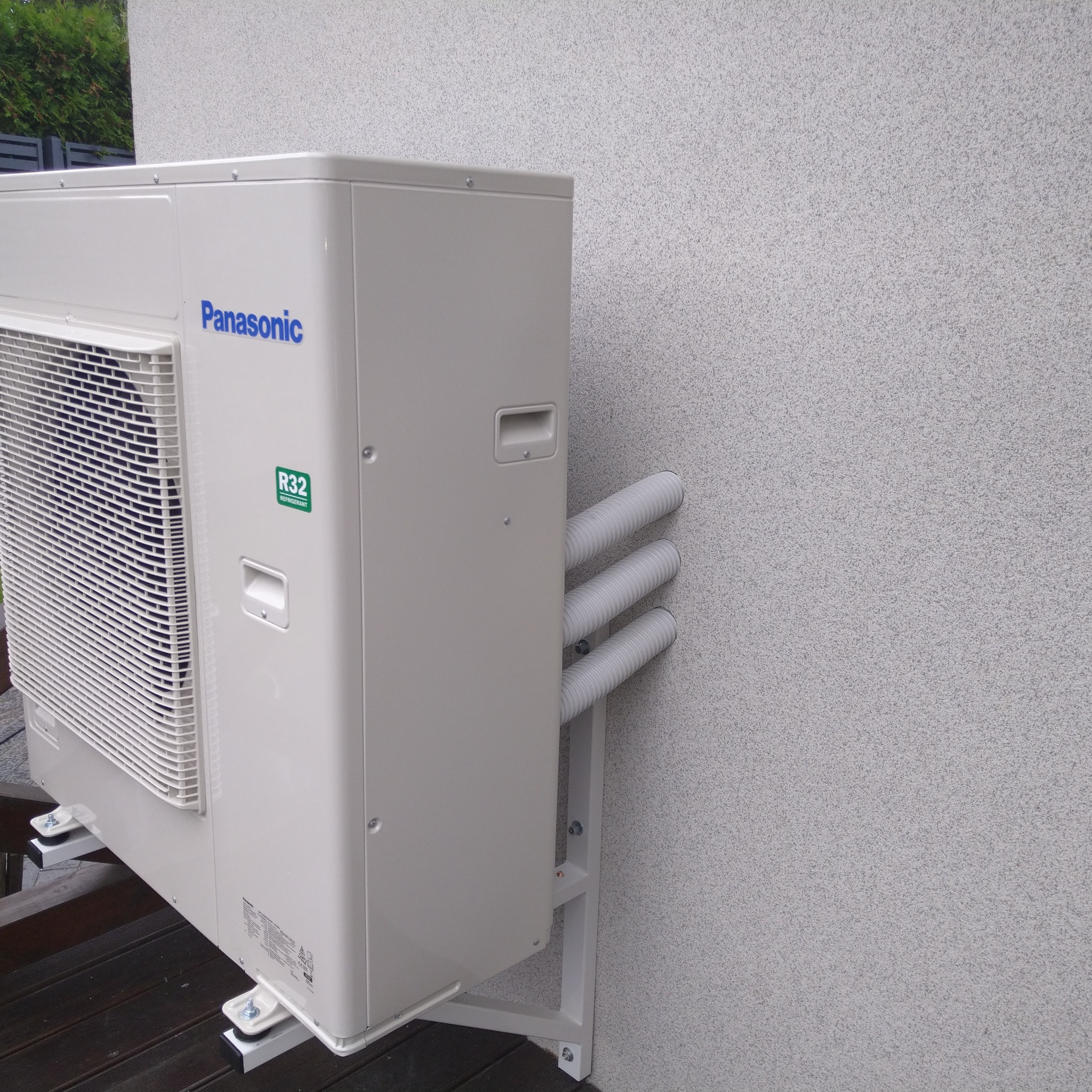Zewnętrzna jednostka klimatyzacji Panasonic zamontowana na ścianie budynku z widocznymi trzema rurami odprowadzającymi, zainstalowana na wsporniku podtrzymującym.