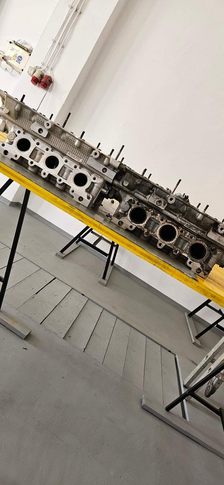 Dwie głowice silnika spalinowego, jedna czysta i druga zabrudzona nagarem, leżące na żółtym stole warsztatowym w jasnym pomieszczeniu.