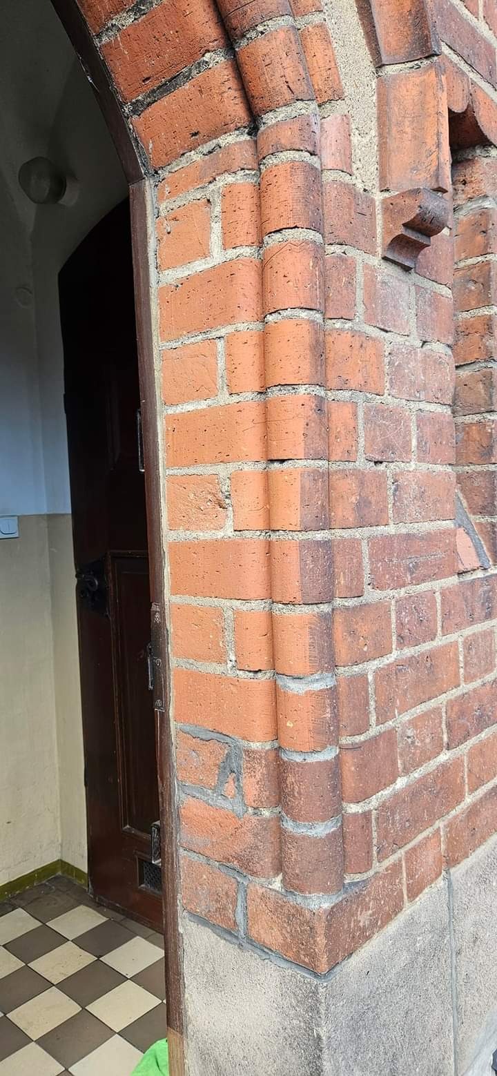 Ceglany portal wejściowy z dekoracyjnymi elementami i widocznym fragmentem drewnianych drzwi oraz szachownicą na podłodze wewnątrz budynku.