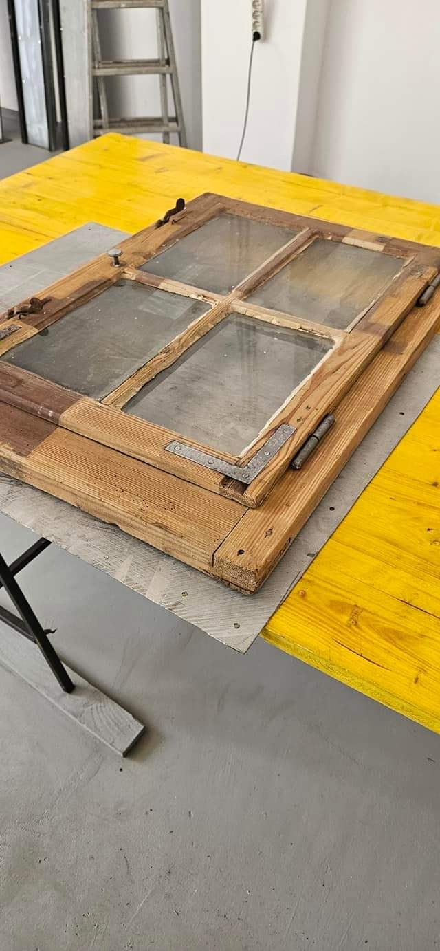 Stara, drewniana rama okienna z szybami, leżąca na żółtym stole roboczym, widoczne metalowe okucia i zawiasy.