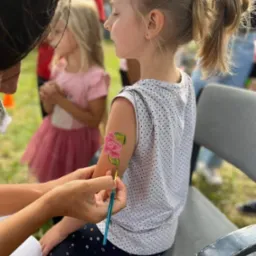 Animator maluje różę na ramieniu dziewczynki siedzącej na krześle, w tle inne dzieci.