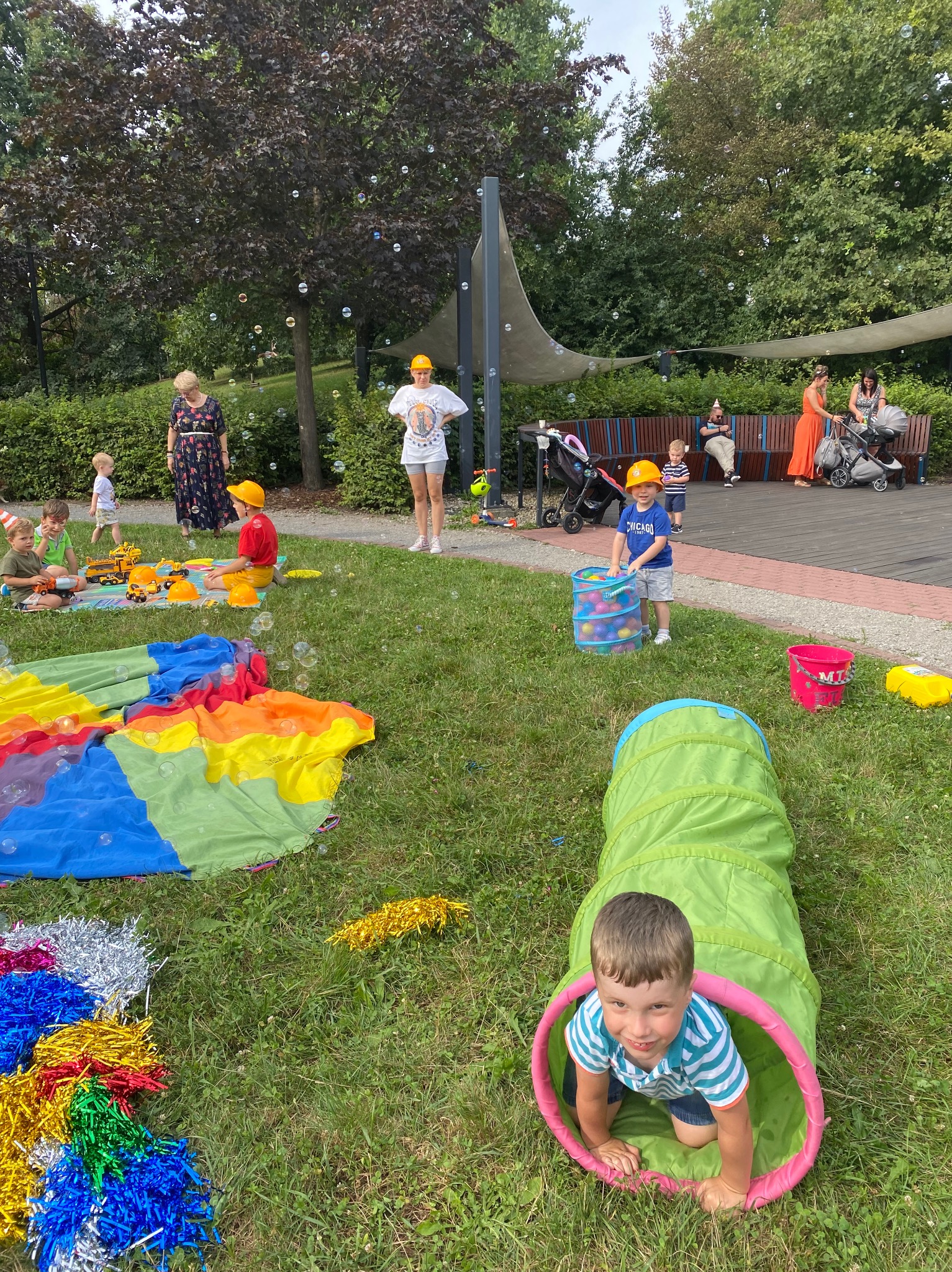 Dzieci bawią się na trawie podczas imprezy plenerowej, w tle dmuchane bańki, tunel, kolorowa chusta animacyjna, wiaderka, piłki i dekoracje.