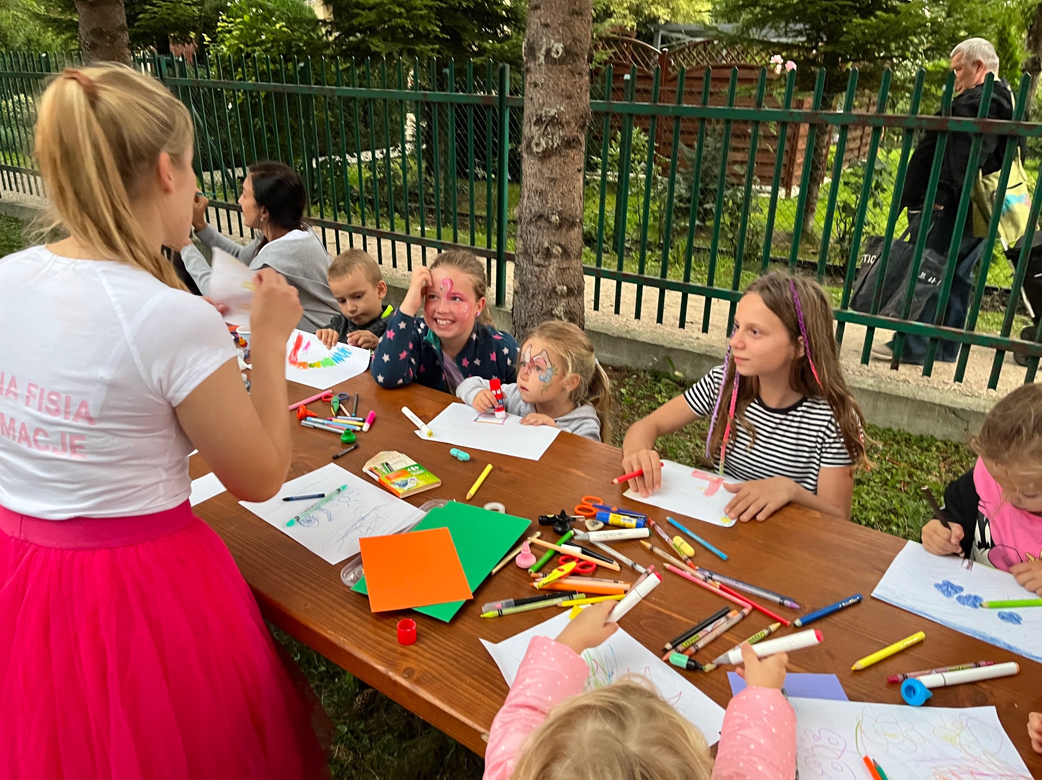 Dzieci siedzące przy drewnianym stole rysują i malują, niektóre mają pomalowane twarze, a animatorka w różowej tiulowej spódnicy nadzoruje zajęcia plastyczne na świeżym powietrzu.