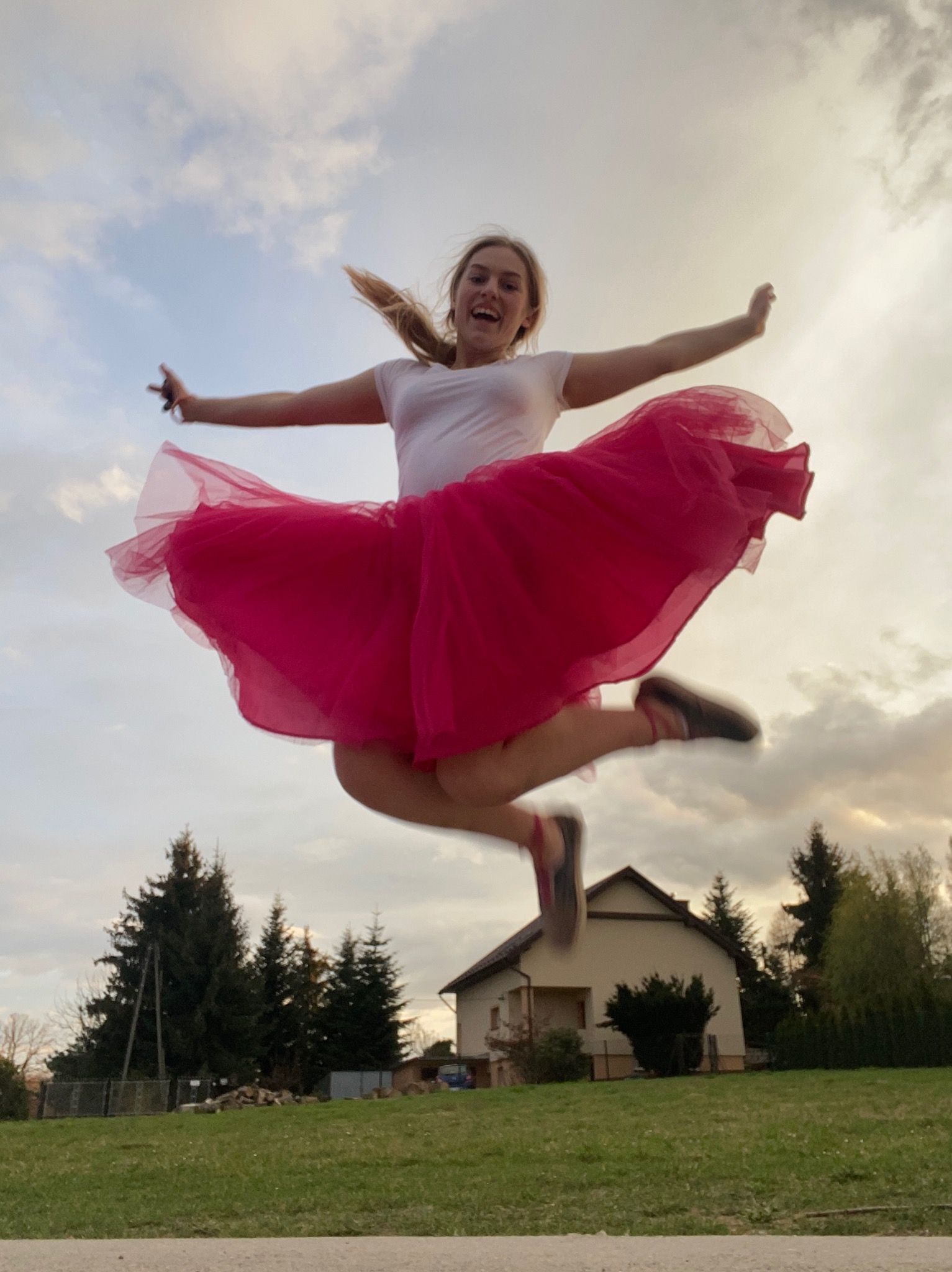 Uśmiechnięta kobieta w białej koszulce i różowej tiulowej spódnicy skacze w powietrzu na tle domu i drzew, z rozpostartymi rękami.