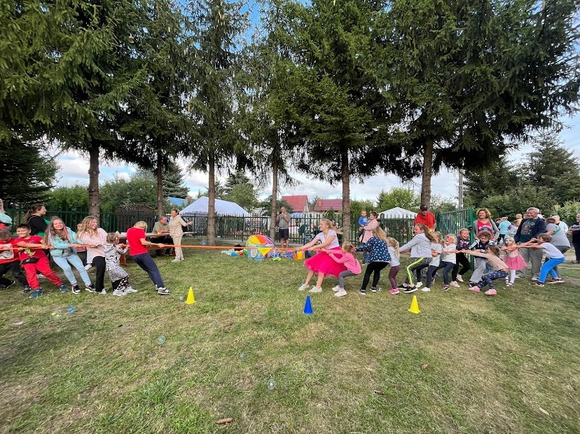 Grupa dzieci i dorosłych bierze udział w przeciąganiu liny na trawiastym terenie podczas imprezy plenerowej, z widocznymi stożkami ograniczającymi pole gry i dmuchanymi bańkami unoszącymi się...