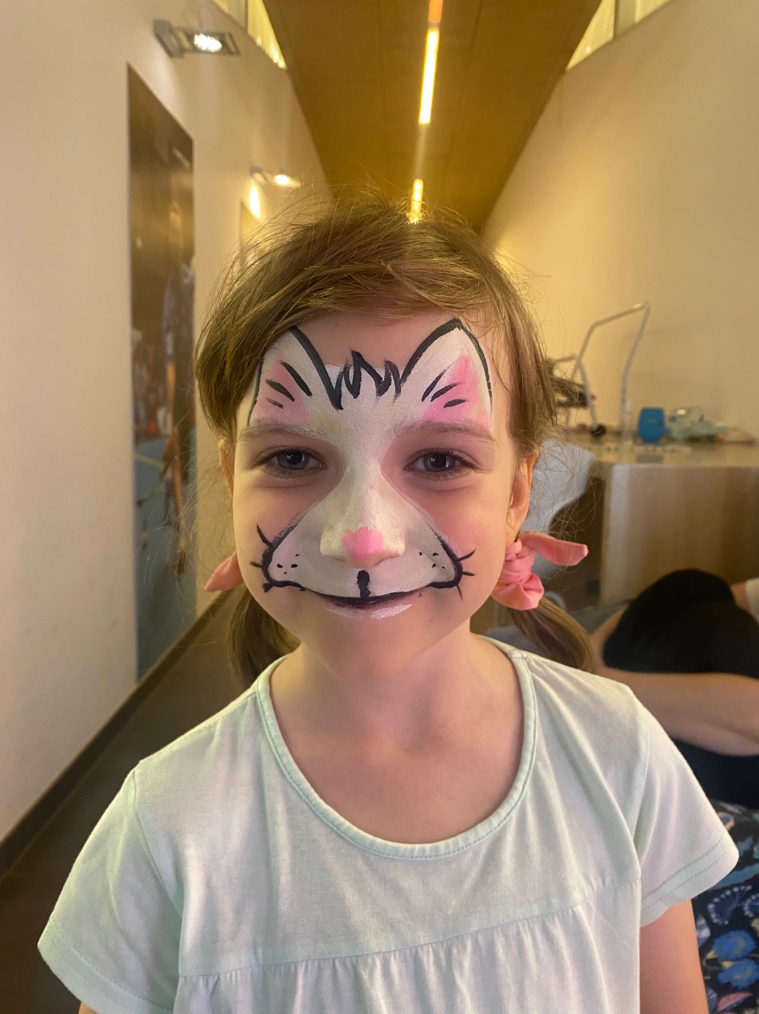 Dziewczynka z pomalowaną twarzą w motywy kota, z różowym nosem i uszami, włosy spięte w kucyki z różowymi gumkami.