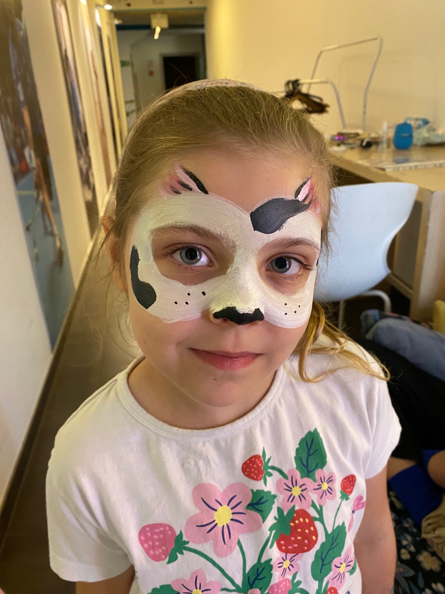 Dziewczynka z pomalowaną twarzą w biało-czarne plamy, przypominające psa, uśmiecha się do kamery, ubrana w białą koszulkę w kwiaty i truskawki.