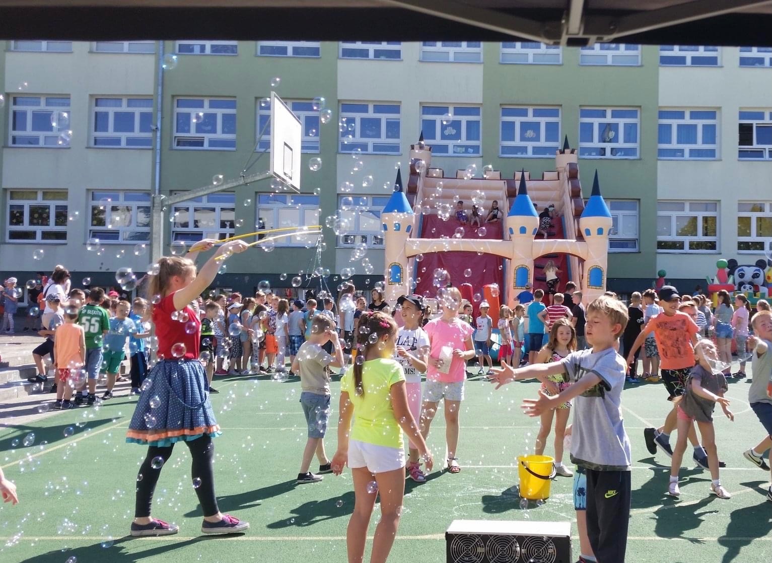 Animator robiący bańki mydlane dla grupy dzieci na tle dmuchanego zamku i budynku szkolnego.