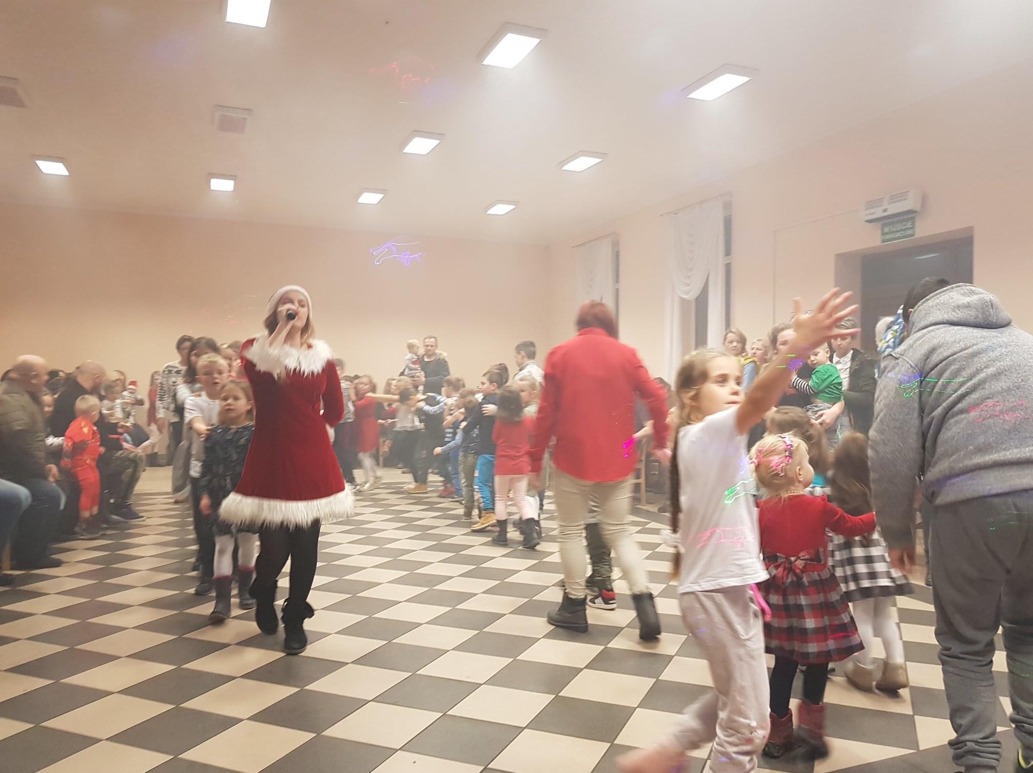 Kobieta w stroju Mikołajki śpiewa do mikrofonu, wokół niej grupa dzieci w różnym wieku podczas imprezy na parkiecie w szachownicę.