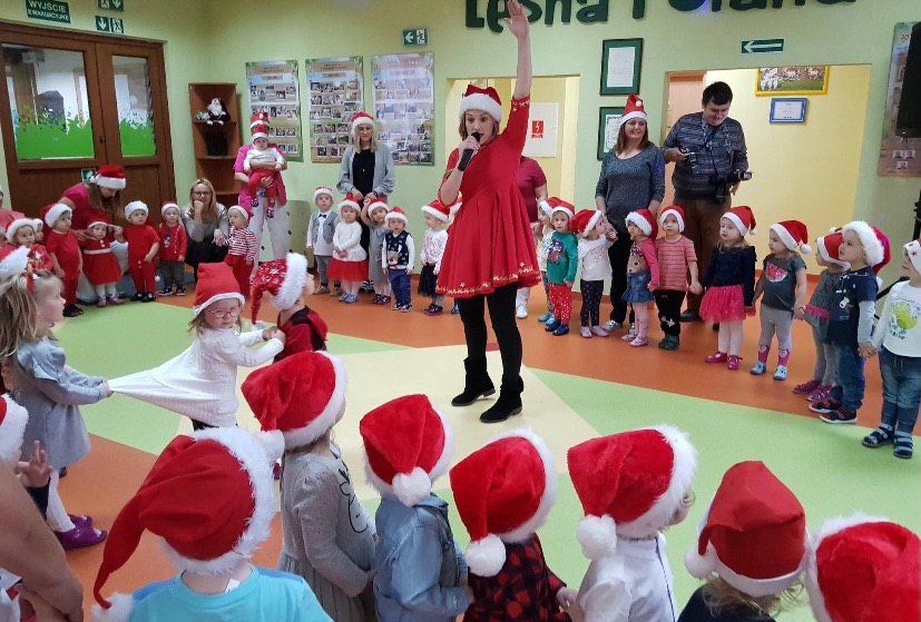 Animator w czerwonej sukience i czapce Mikołaja, śpiewa do mikrofonu, otoczona grupą dzieci w czapkach Mikołaja podczas zabawy w przedszkolu, w tle rodzice i dekoracje świąteczne.