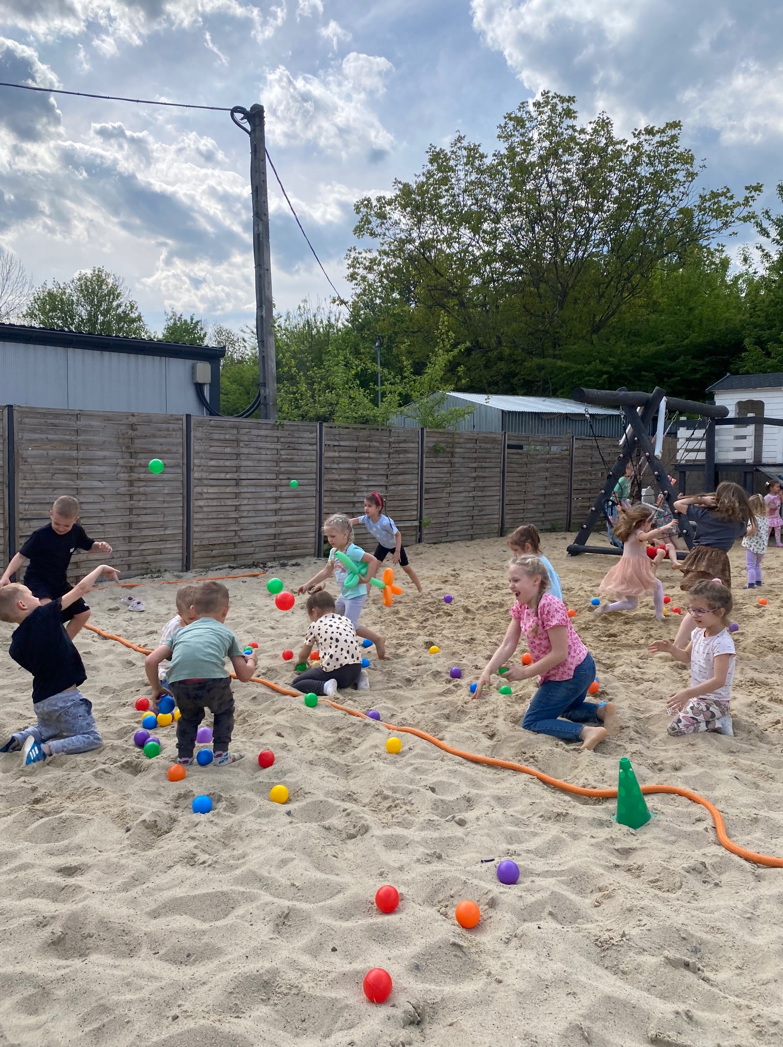 Grupa dzieci bawi się kolorowymi piłkami w piaskownicy na świeżym powietrzu, w tle drewniany płot, huśtawka i słup elektryczny.