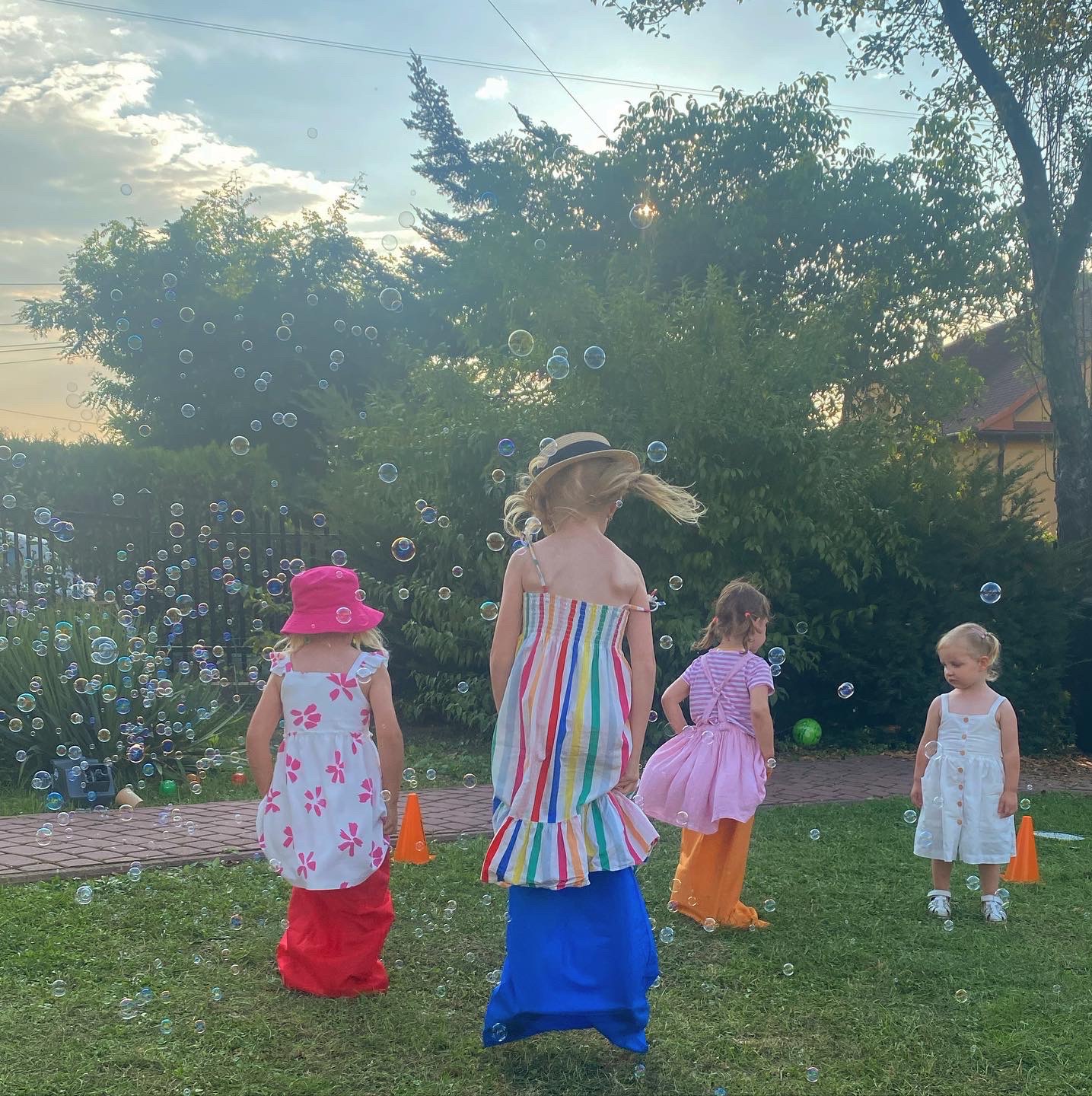 Cztery dziewczynki w kolorowych sukienkach bawią się na trawie w otoczeniu baniek mydlanych, ustawione są pomarańczowe pachołki.