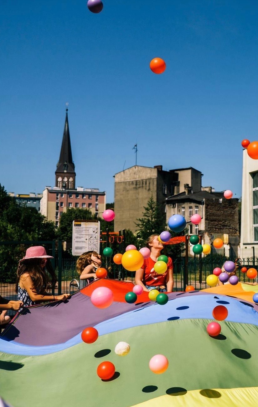 Dzieci i dorosły bawią się kolorową chustą animacyjną, podrzucając w górę różnokolorowe piłeczki na tle miejskiego krajobrazu z kościelną wieżą.