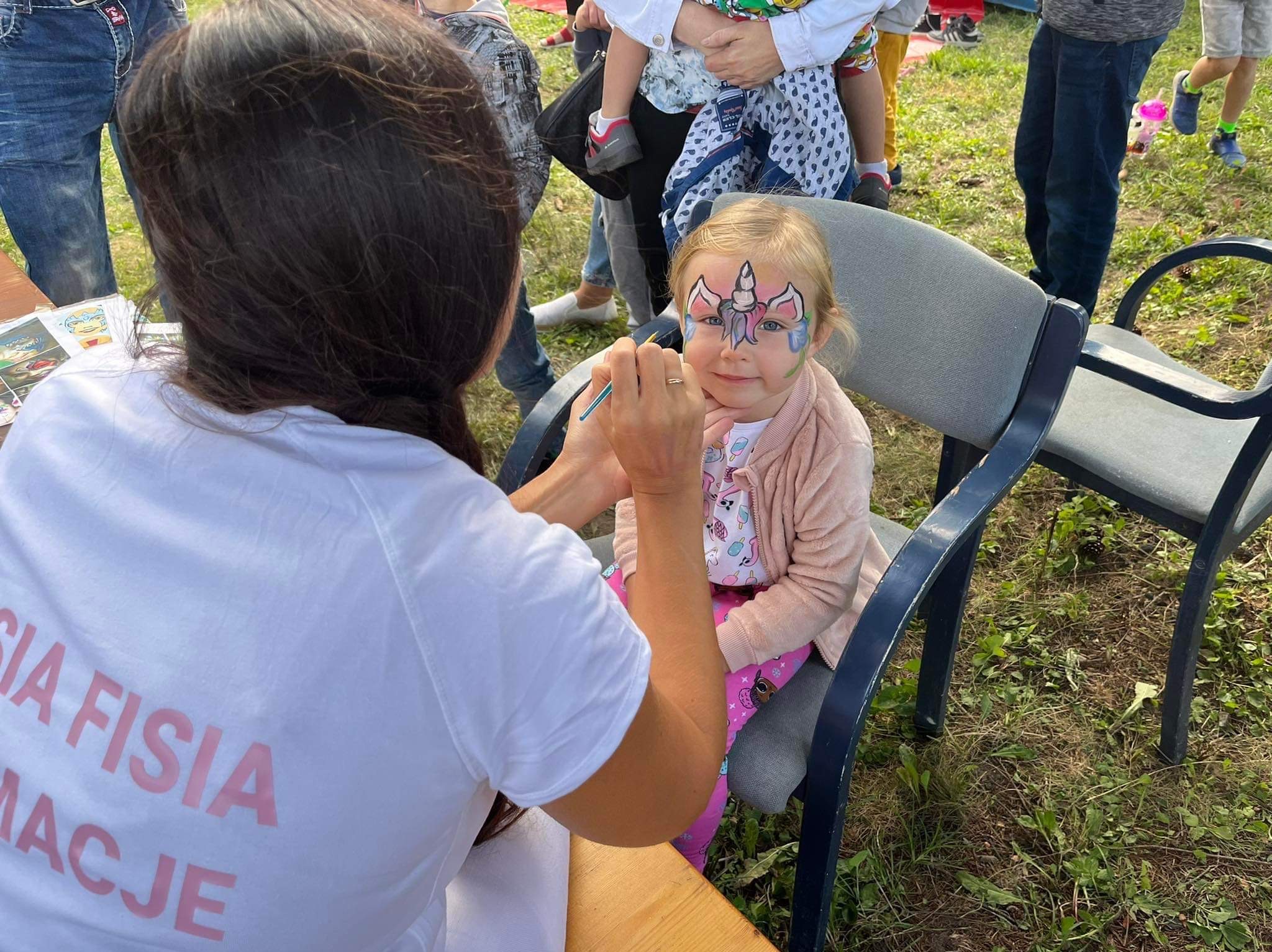 Dziewczynka z pomalowaną twarzą w jednorożca siedzi na krześle, podczas gdy artystka maluje jej policzek pędzlem, w tle widoczni ludzie i trawa.