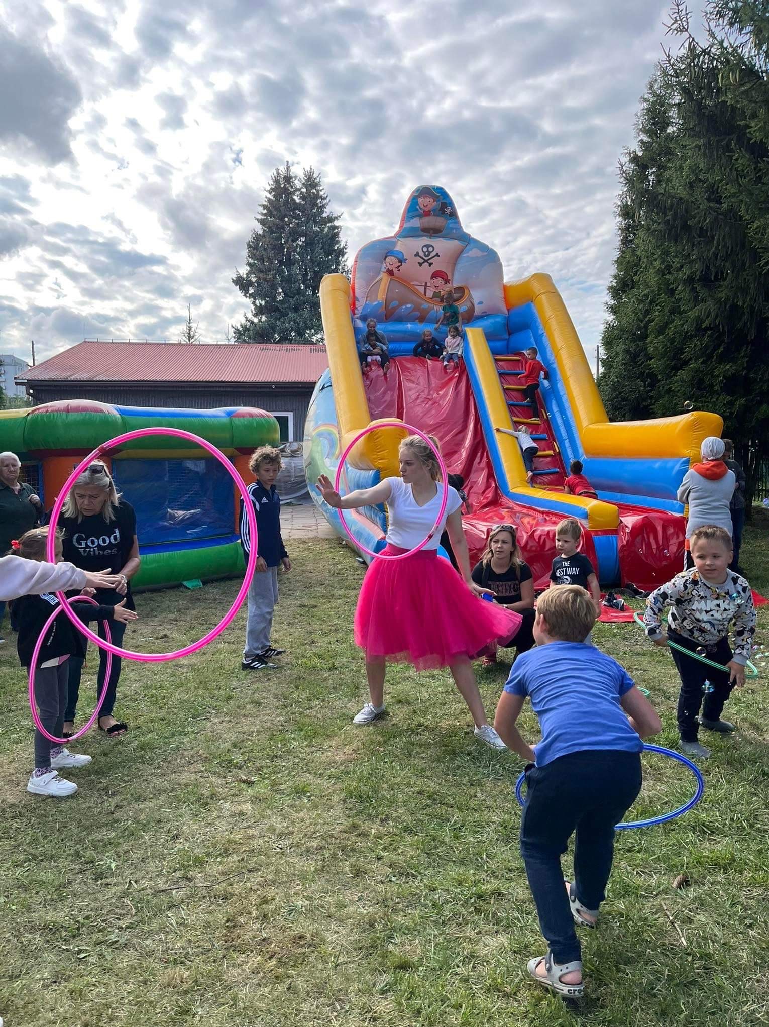 Dzieci bawią się na trawie z kolorowymi obręczami hula-hoop, w tle dmuchany zamek w kształcie pirata, na którym bawią się inne dzieci.