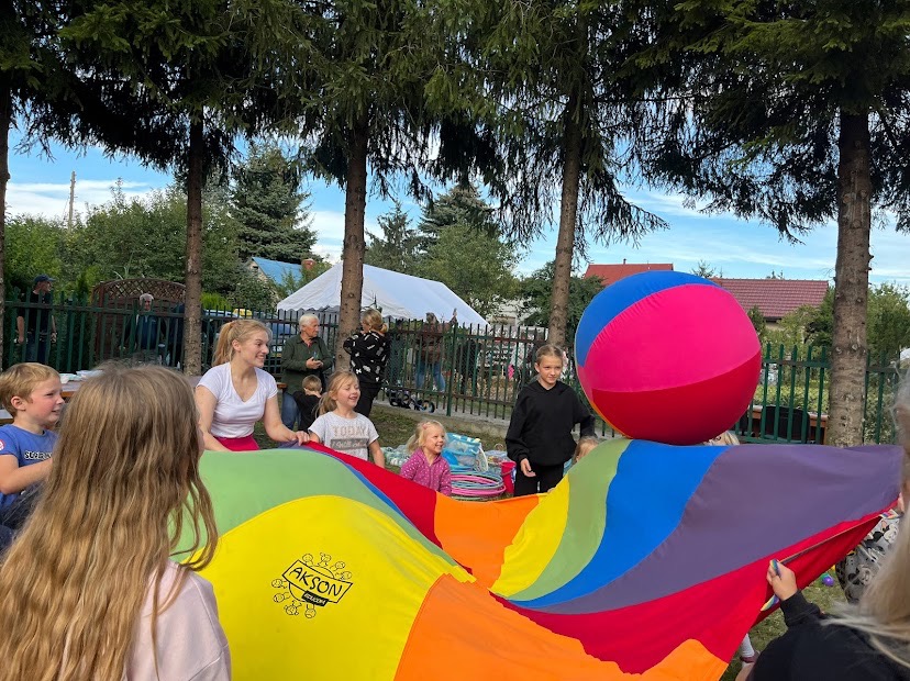 Grupa dzieci i dorosłych bawi się kolorową chustą animacyjną z dużym, trójkolorowym balonem na wierzchu, na tle zieleni i białego namiotu.