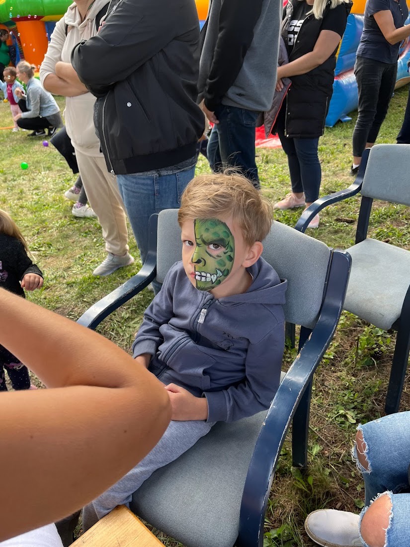 Chłopiec z pomalowaną połową twarzy na zielono w motywie jaszczurki siedzi na krześle na tle dmuchanego zamku i innych dzieci.