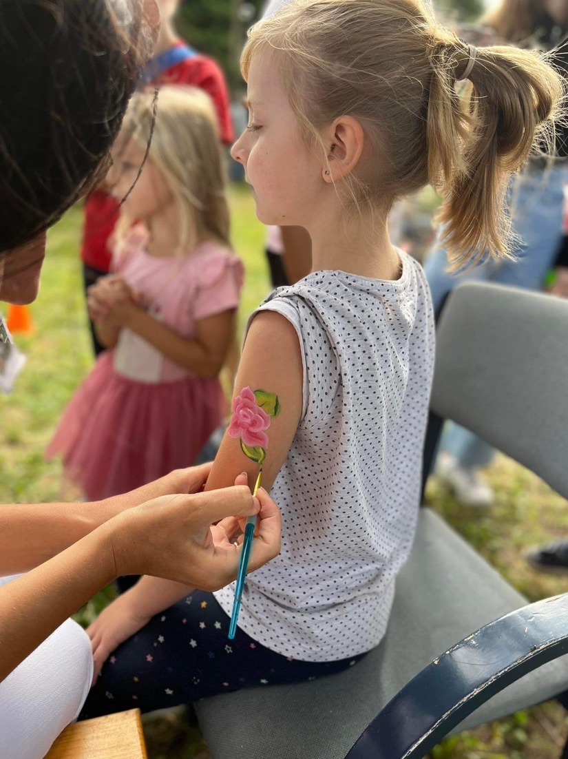 Animator maluje różę na ramieniu dziewczynki siedzącej na krześle, w tle inne dzieci.