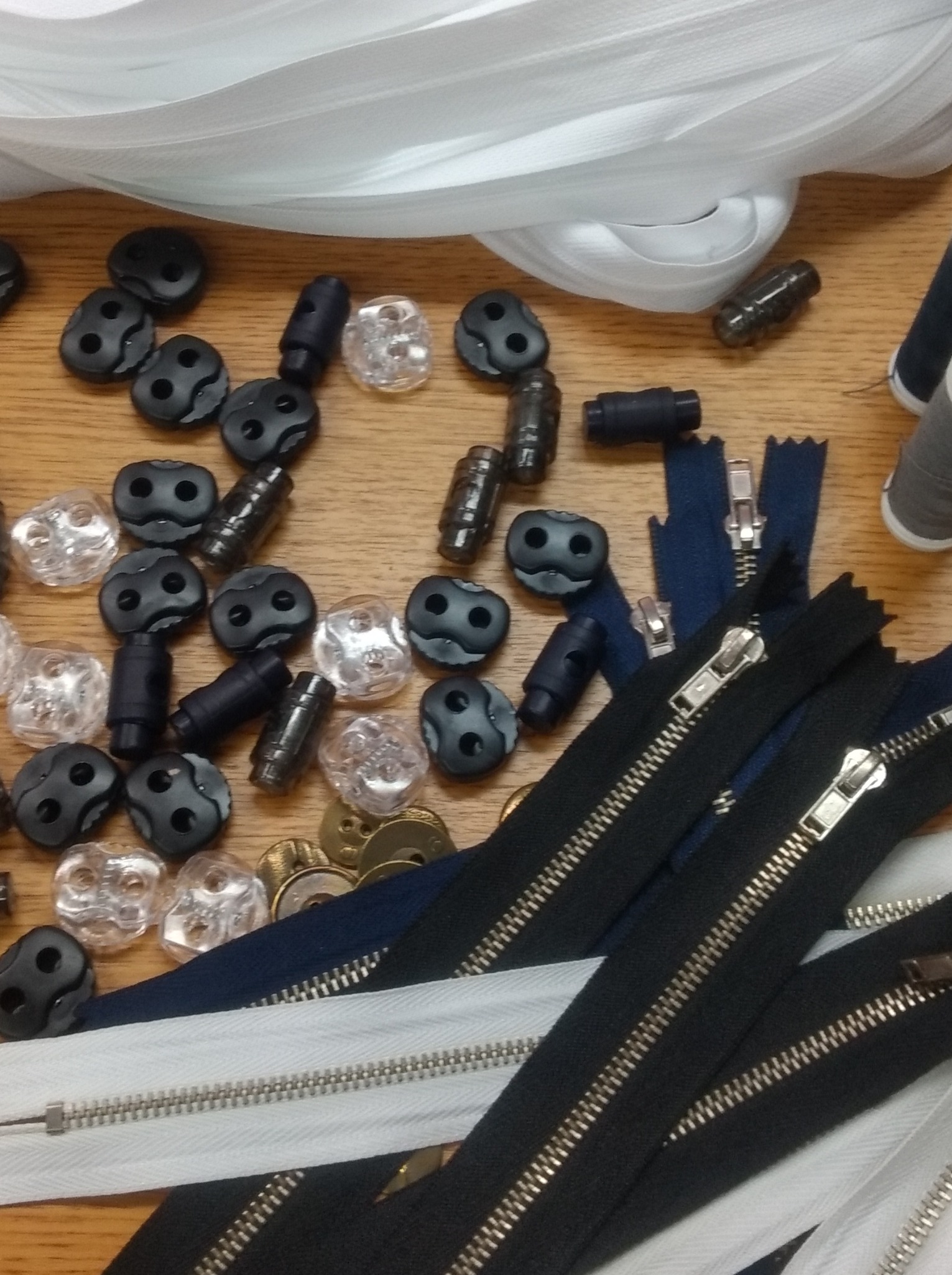 Różnorodne zamki błyskawiczne w kolorach czarnym i białym, okrągłe czarne i przezroczyste stopery, guziki i nici na drewnianym stole, widoczne z góry.