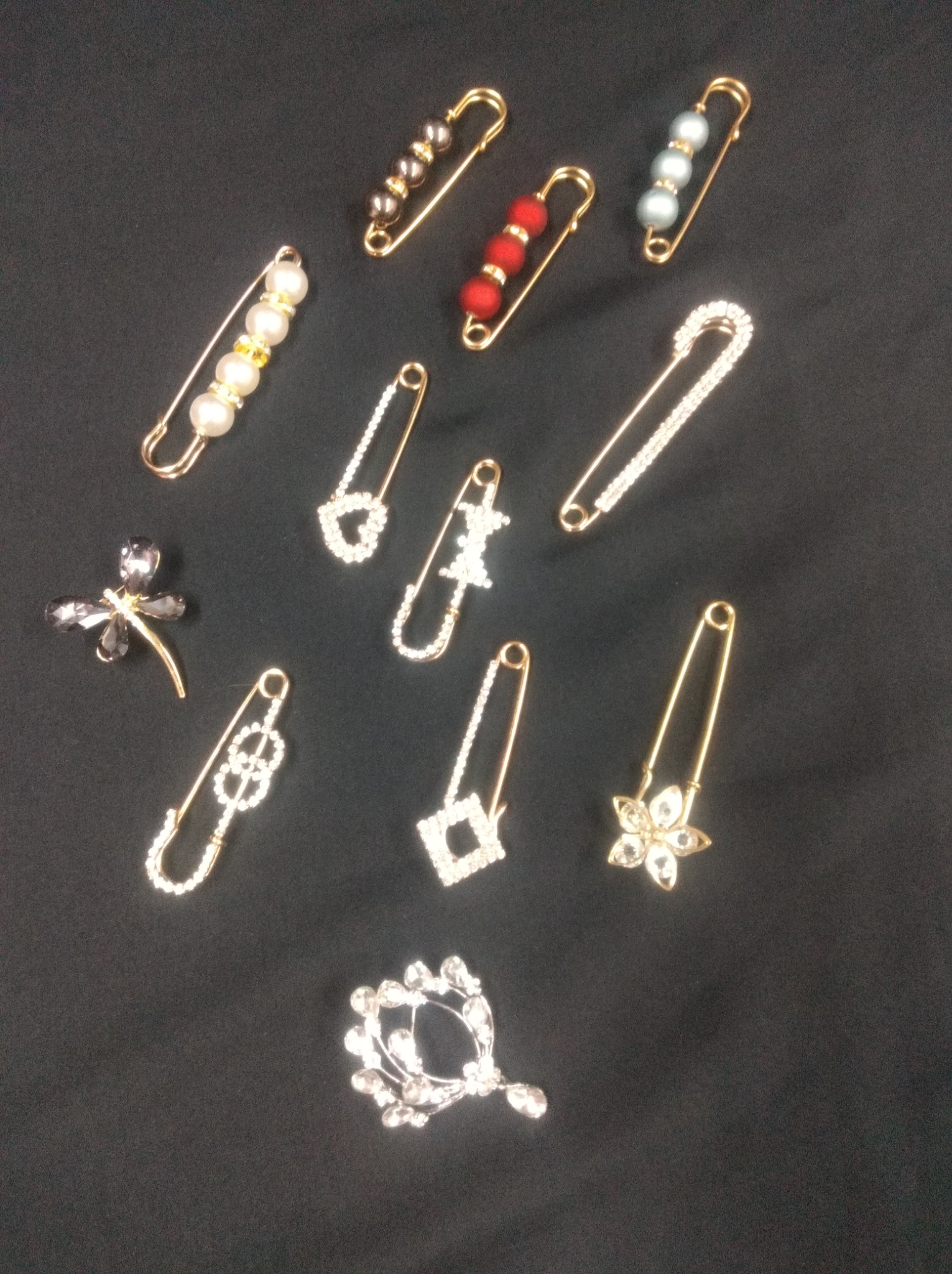 Zestaw ozdobnych agrafek i broszek z koralikami i cyrkoniami w różnych wzorach, ułożonych na czarnym tle.