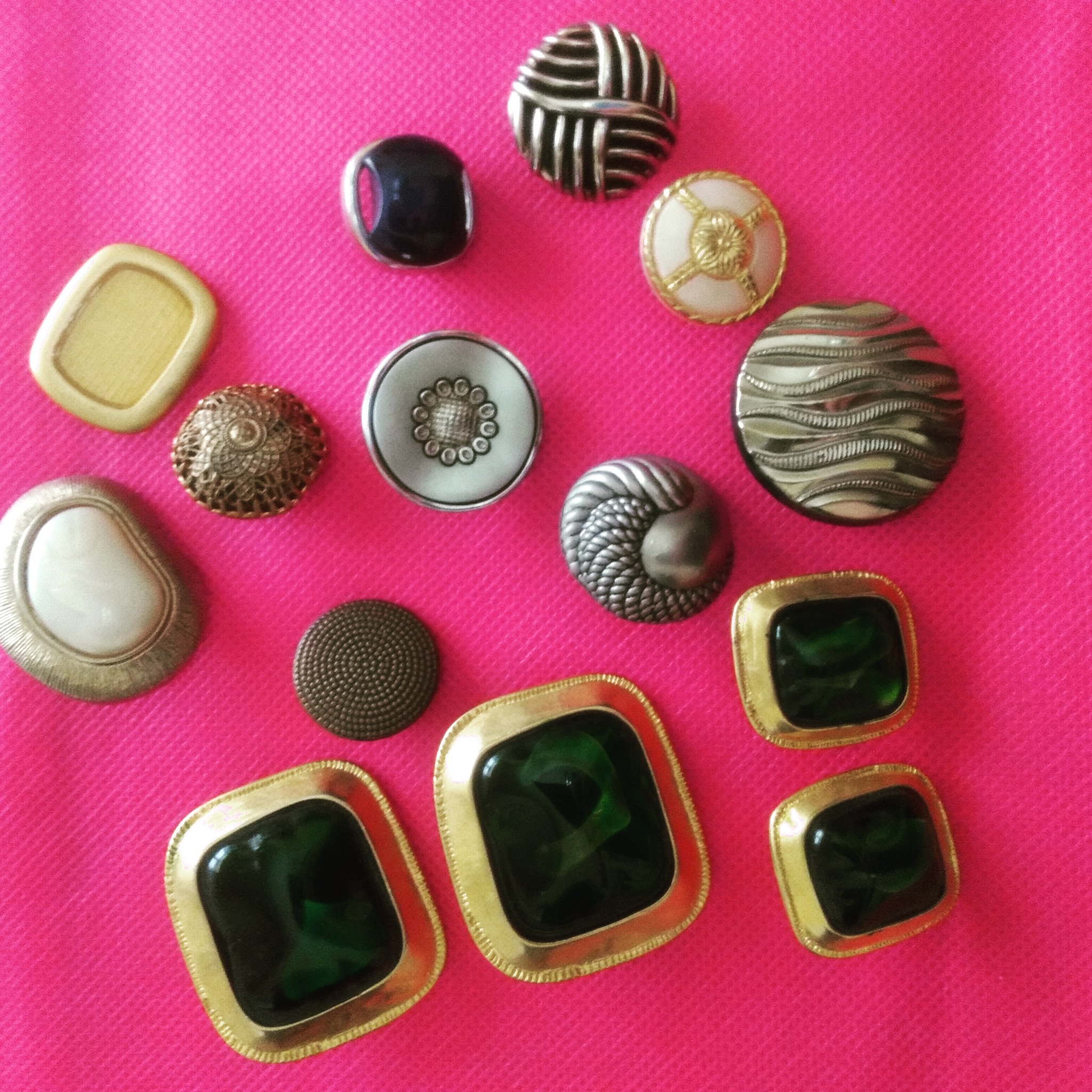 Zestaw ozdobnych guzików o różnych kształtach, kolorach i fakturach na jaskraworóżowym tle materiału o widocznej fakturze. Guziki metalowe, plastikowe, z zielonymi wstawkami, okrągłe i kwadratowe...