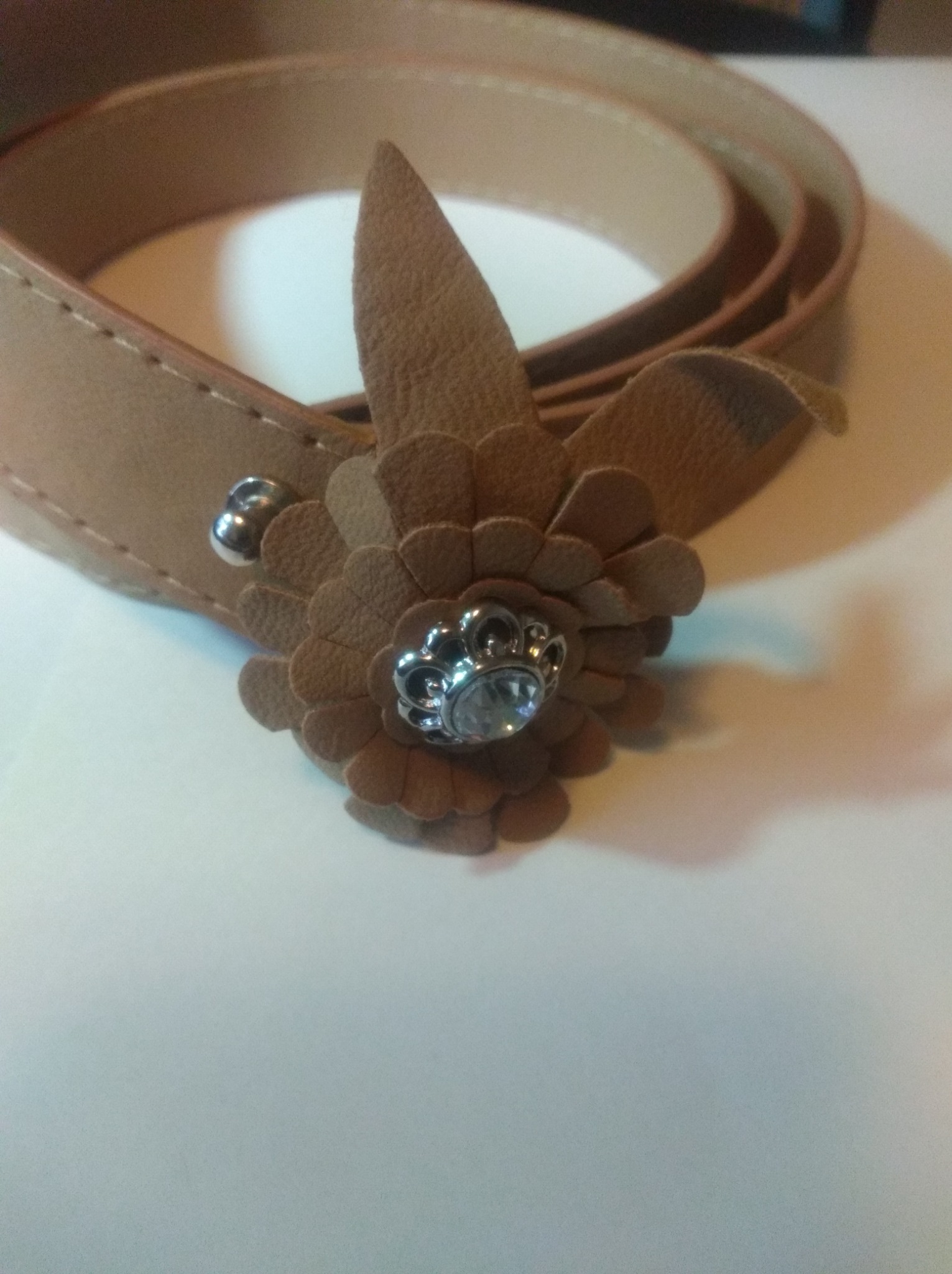 Beżowy, skórzany pasek damski z ozdobną, brązową aplikacją w kształcie kwiatu z błyszczącym, srebrnym elementem pośrodku, leżący na białym tle.