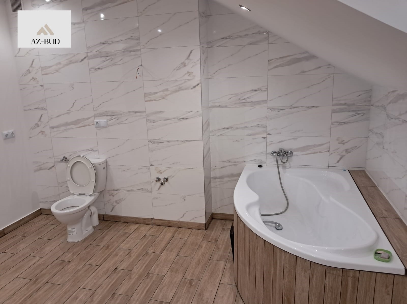 Wykończona łazienka na poddaszu z białą wanną narożną obudowaną drewnem, toaletą i marmurkowymi płytkami na ścianach, widoczny logotyp firmy w górnym rogu.